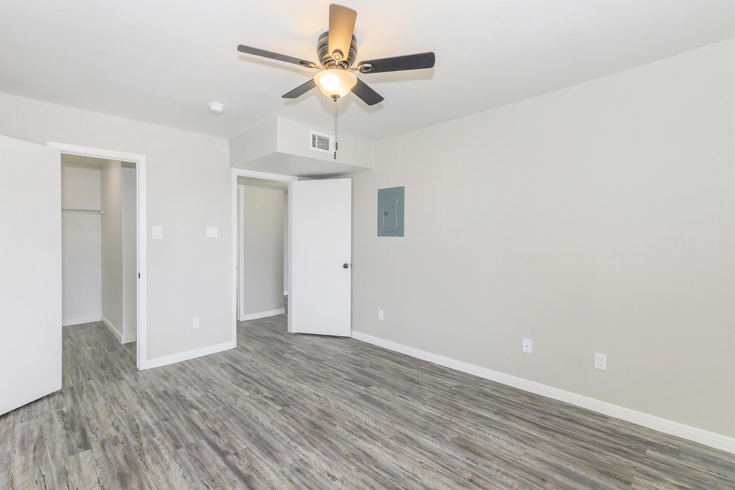 Spacious, modern room with light gray walls and laminate flooring. Features a ceiling fan, two open doors leading to closets, and a small electrical panel on the wall. Natural light fills the space, creating a bright and inviting atmosphere. Ideal for a bedroom or study.