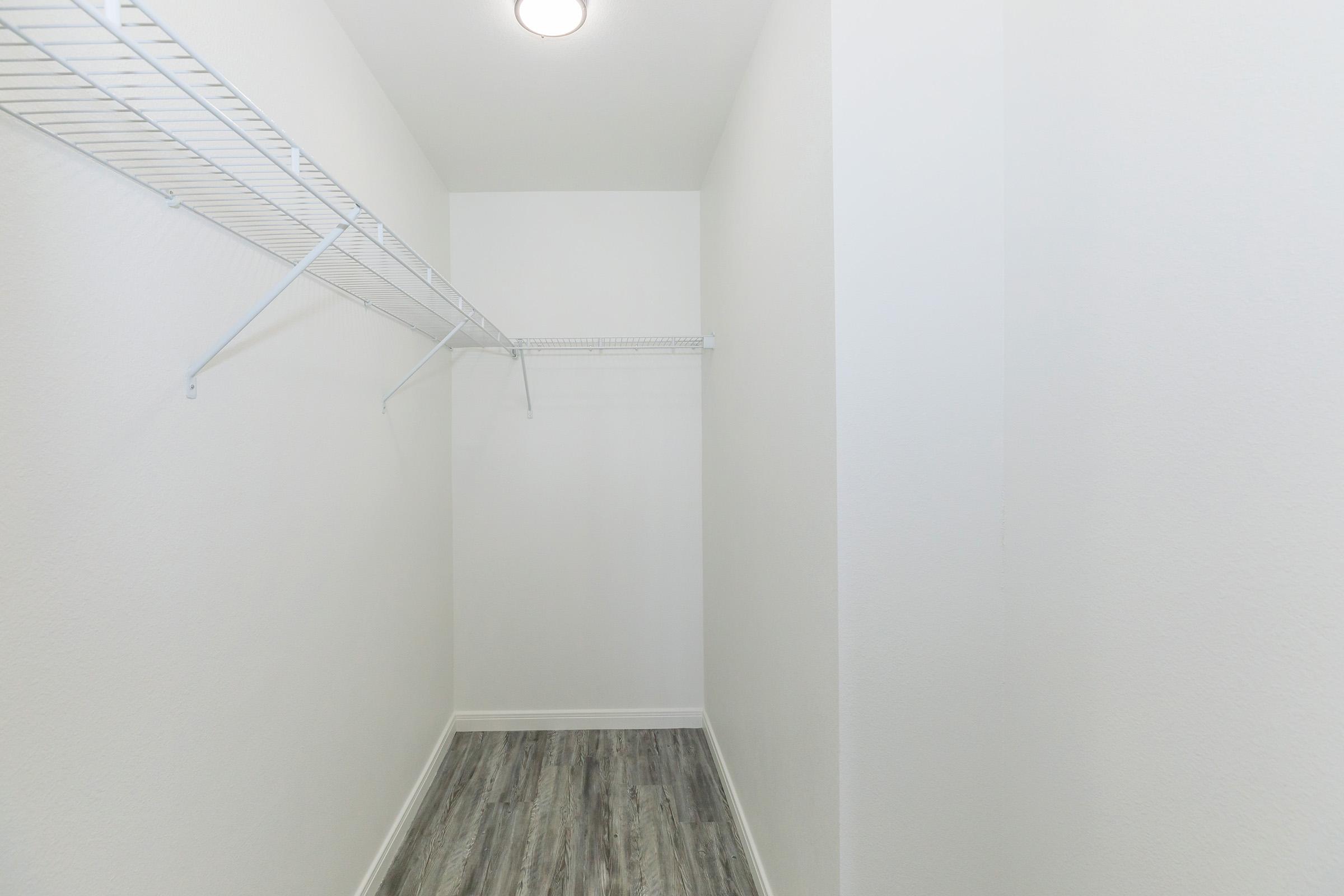 A clean, empty closet featuring white walls, a light-colored wooden floor, and a wire shelving unit along one side. The minimalist design gives a sense of spaciousness, with plenty of natural light illuminating the area.