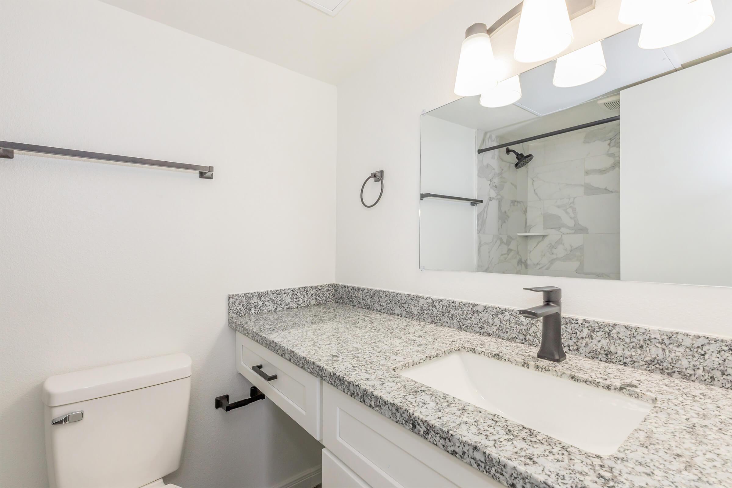 A modern bathroom featuring a granite countertop with a rectangular sink, a wall-mounted faucet, a mirror above, and a shower area in the background. The walls are painted white, and there is a toilet positioned beside the sink. Soft lighting is provided by three pendant lights above the mirror.