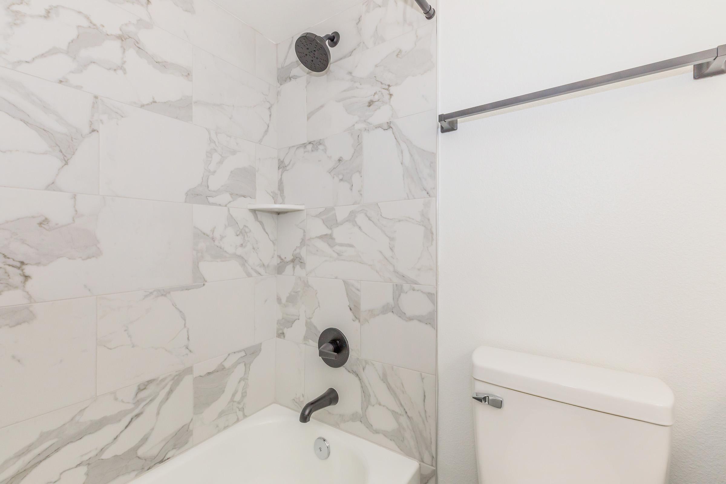 A modern bathroom featuring a white bathtub with a marble tile shower wall. The shower has a stylish overhead showerhead and a wall-mounted faucet. There’s a small shelf in the shower for toiletries and a white toilet adjacent to the bath area. The overall design is contemporary and clean.