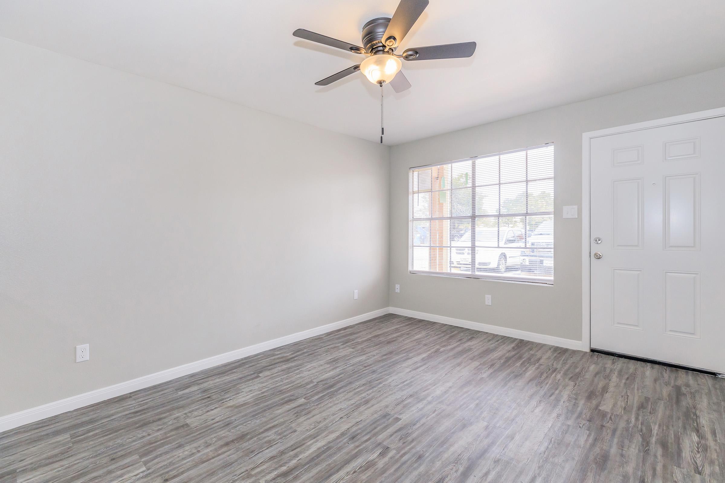 A bright and spacious room with light gray walls, a ceiling fan, and new wood-look flooring. A large window allows natural light to fill the space, and there's a front door visible on the right side. The room is empty, offering a clean slate for furnishing.