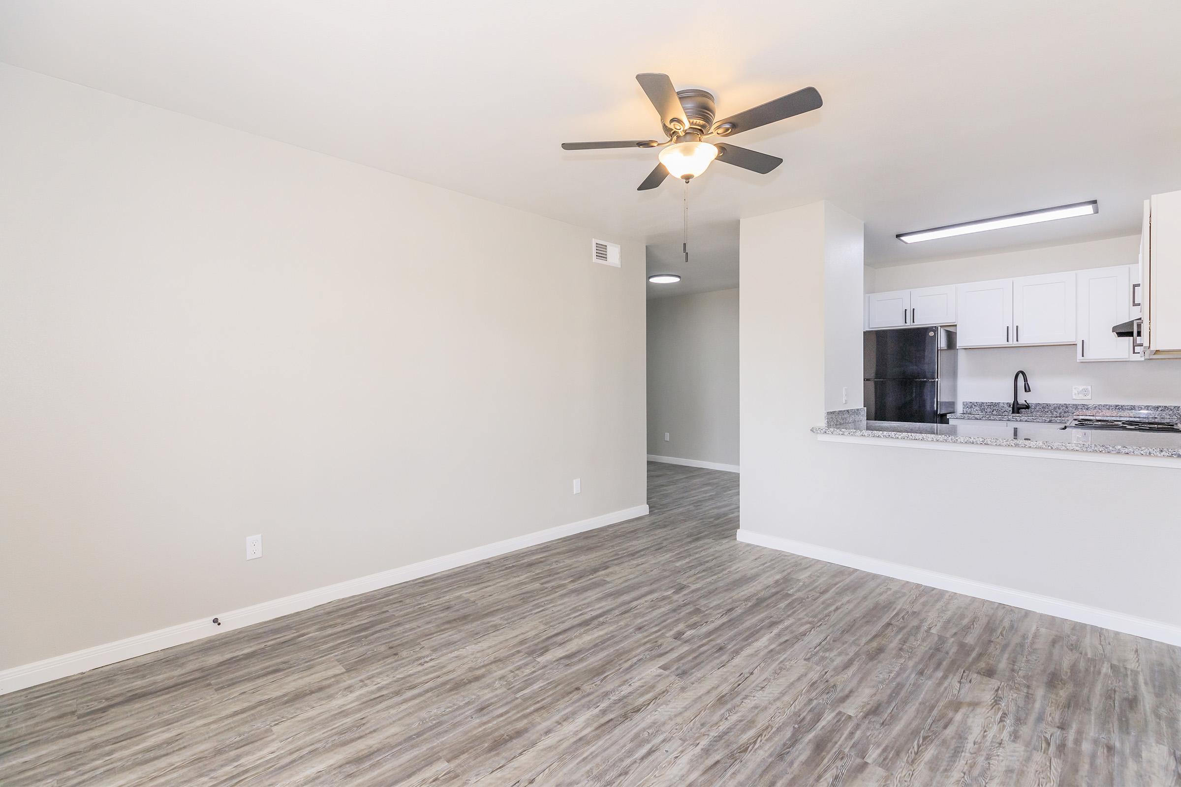 Spacious interior of a modern apartment featuring light-colored walls, a ceiling fan, and laminate flooring. The open layout connects the living area to a kitchen with white cabinets and granite countertops. There's a doorway leading to another room in the background.