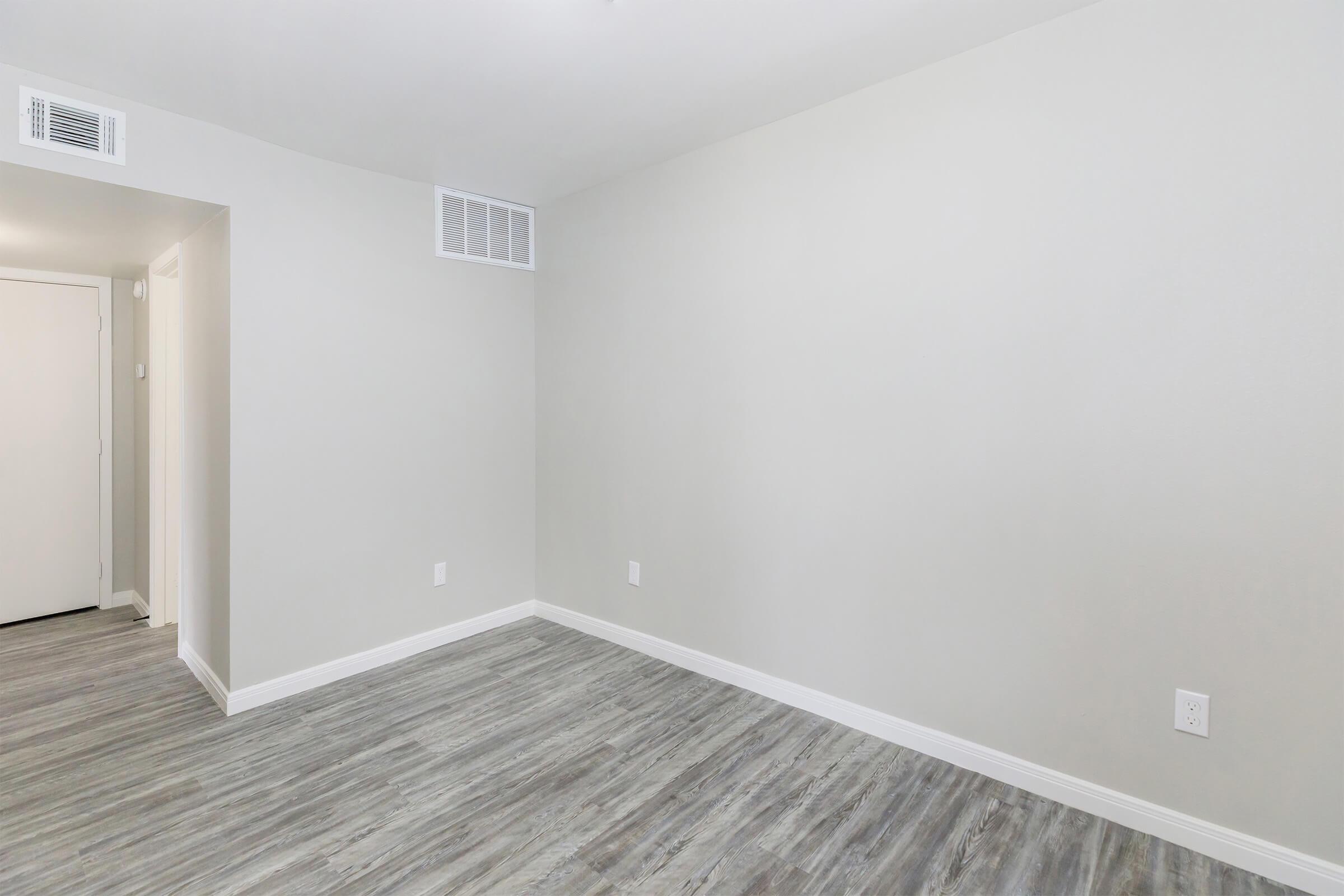 A clean, empty room with light gray walls and light wooden flooring. One wall features an air vent, and there is a door to the left, suggesting an entrance. The overall atmosphere is minimalistic and inviting.