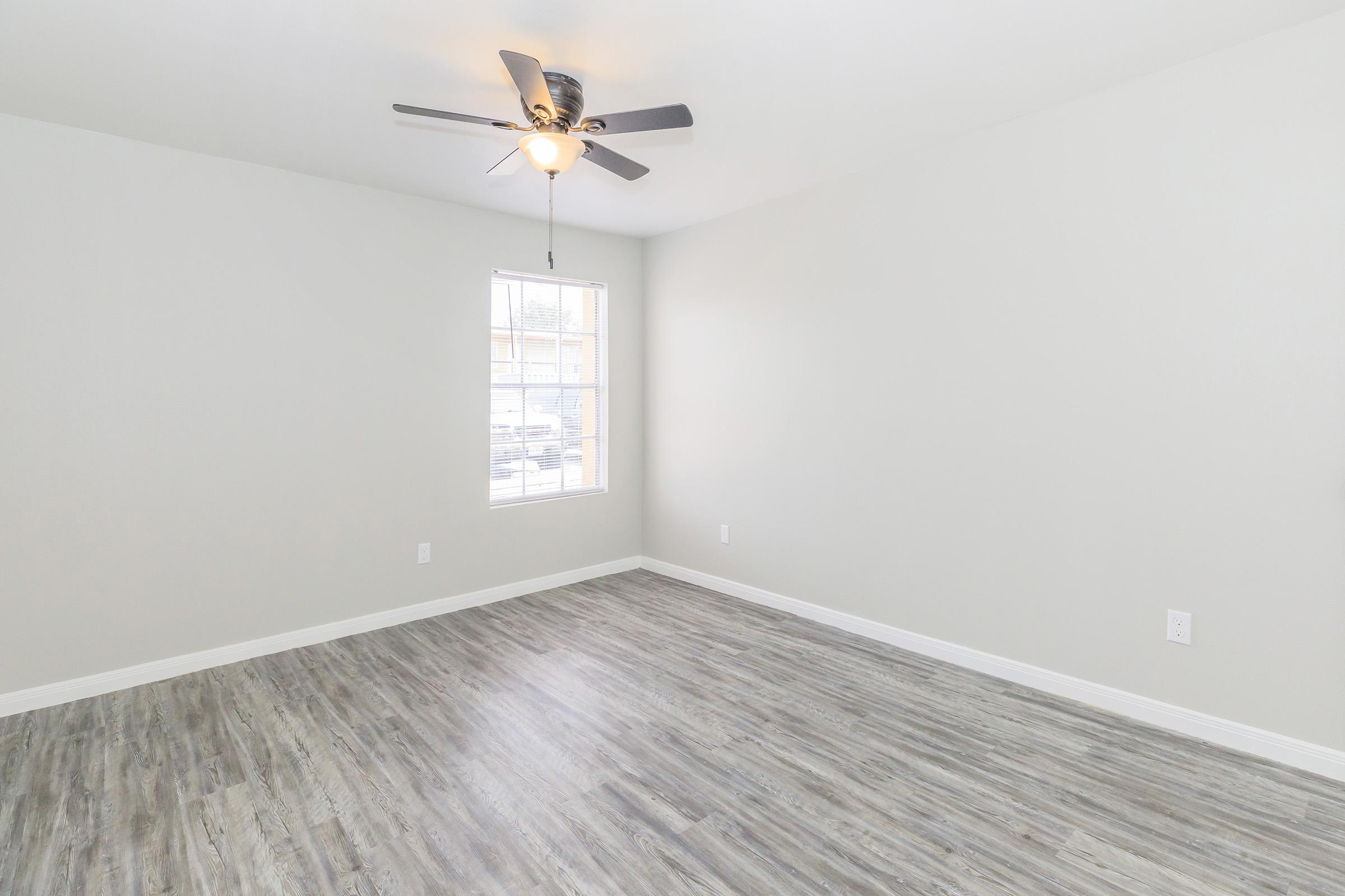 An empty room with light gray walls and a ceiling fan. The floor features light wood-like tiles. A single window allows natural light to enter, but the space is otherwise unadorned, creating a minimalist aesthetic.