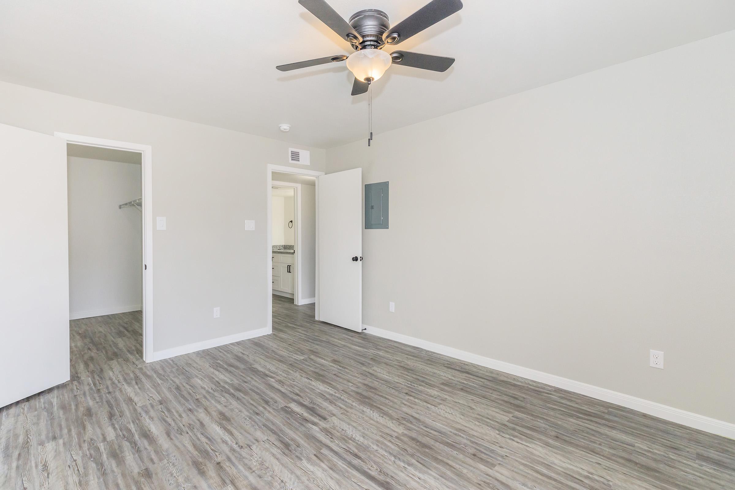 A spacious, empty room featuring light-colored walls, a ceiling fan with black blades, and laminate flooring. Two white doors lead to adjacent areas, with one door partially open, revealing a glimpse of a bathroom or utility area. Natural light brightens the room, enhancing the modern, clean aesthetic.