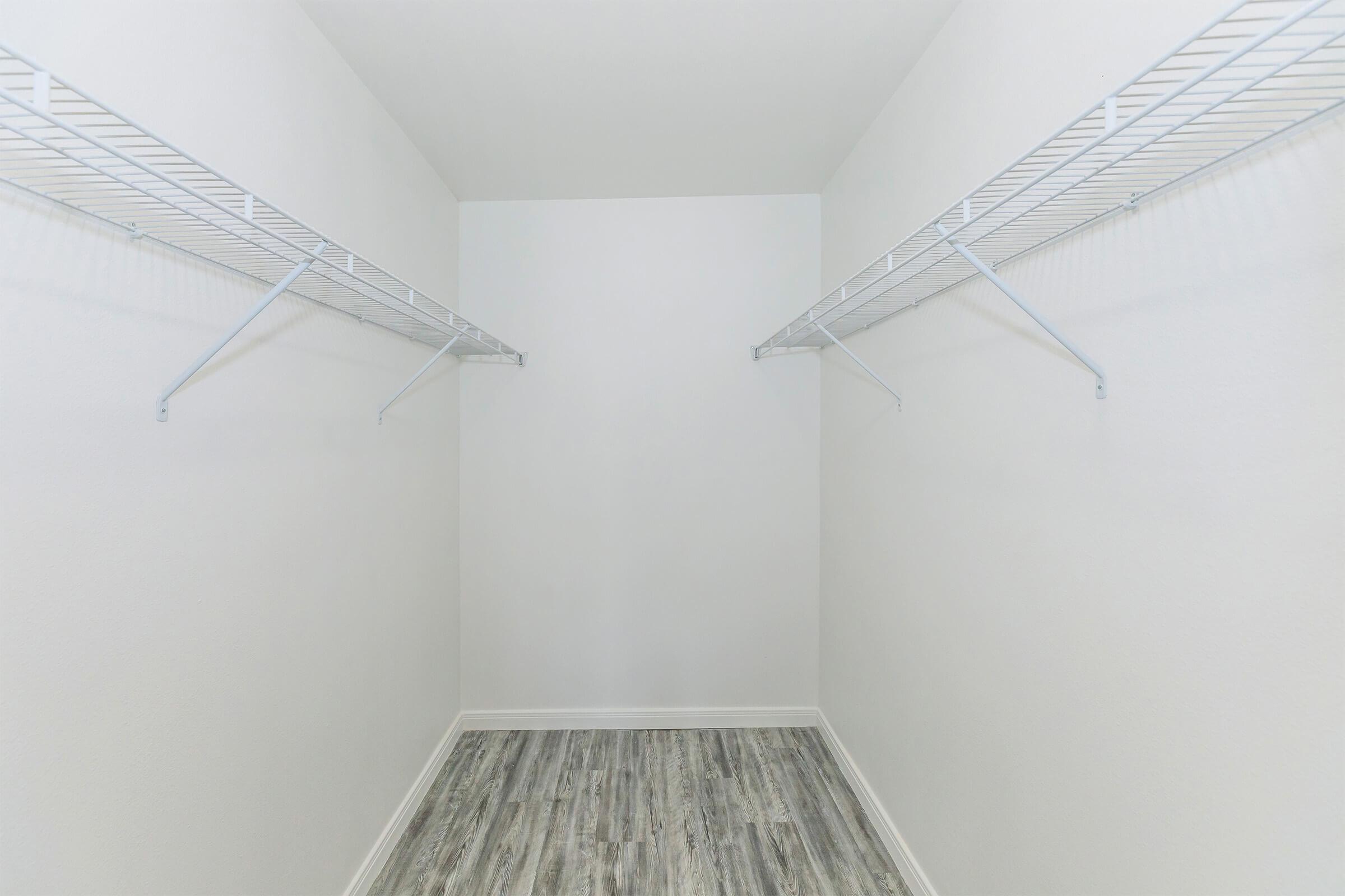 Empty closet with white walls and a light wood floor. Two wire shelves are mounted on opposite walls, providing storage space. The overall appearance is clean and minimalistic, emphasizing the spaciousness of the area.