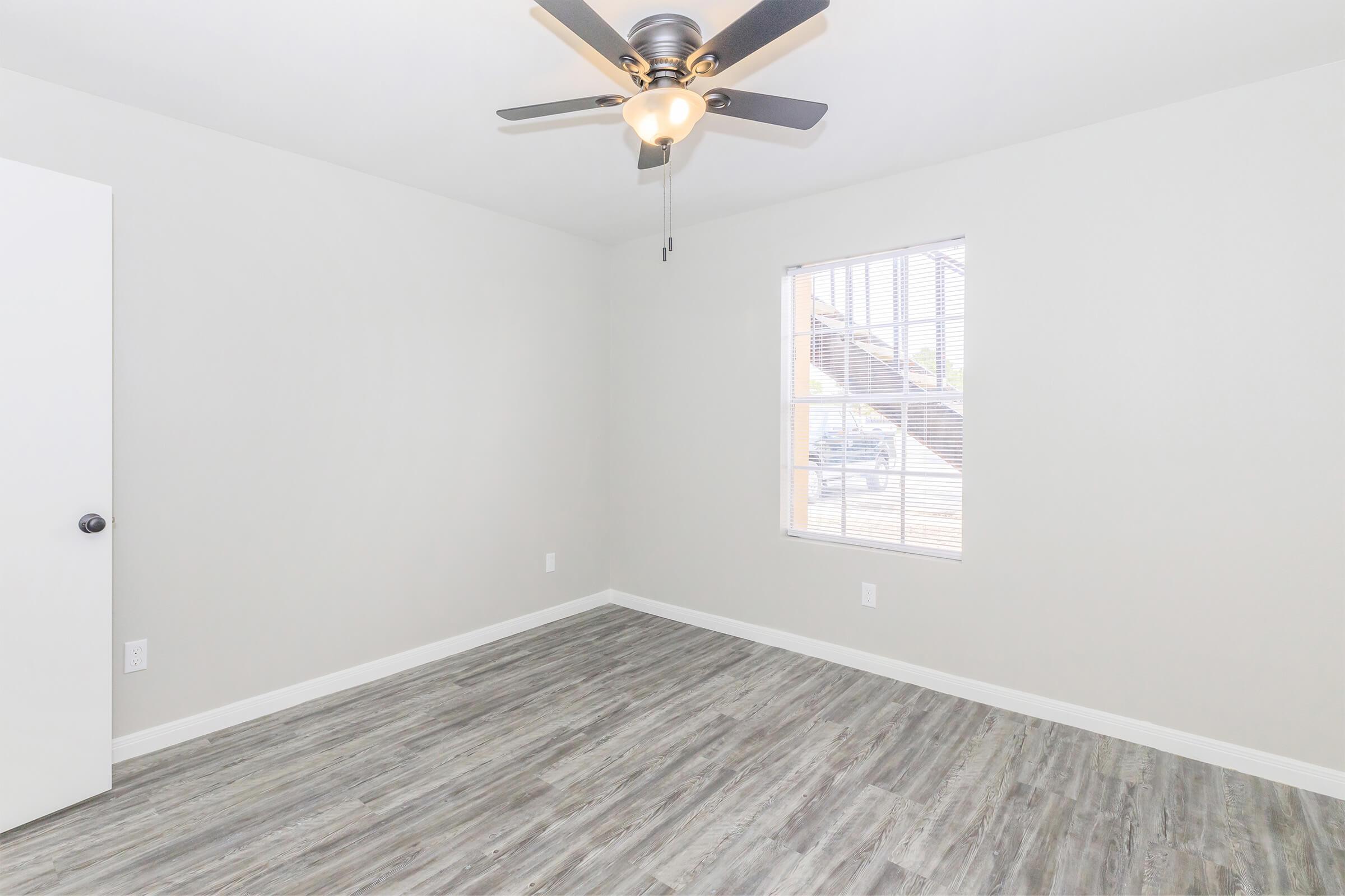 A bright, empty room with light gray walls and a ceiling fan. The floor is a light wood-like laminate. A window allows natural light to enter, showcasing vertical blinds. An open white door leads to another area, enhancing the spacious feel of the room.