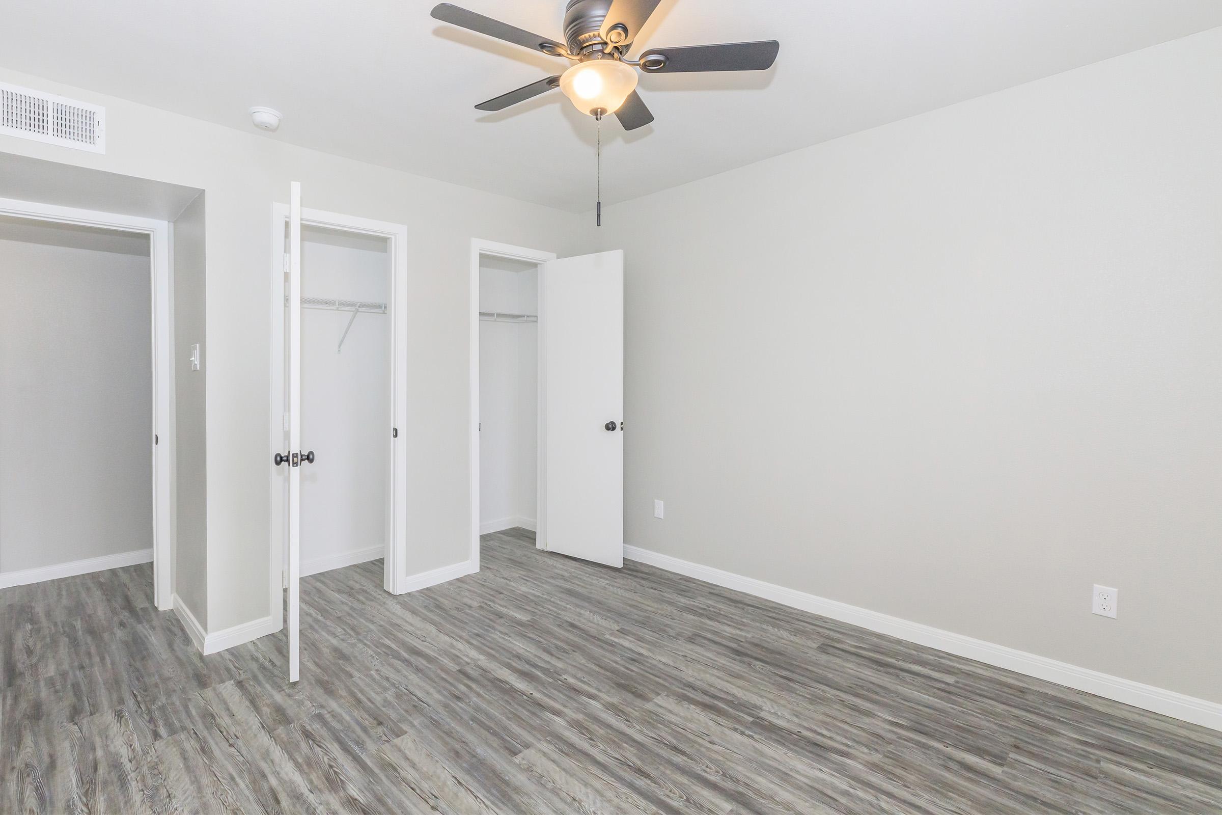 A modern and spacious bedroom featuring light gray walls, wood-like flooring, a ceiling fan, and two open closet doors. The room is empty, providing a clean and minimalist appearance, with natural light flowing in.