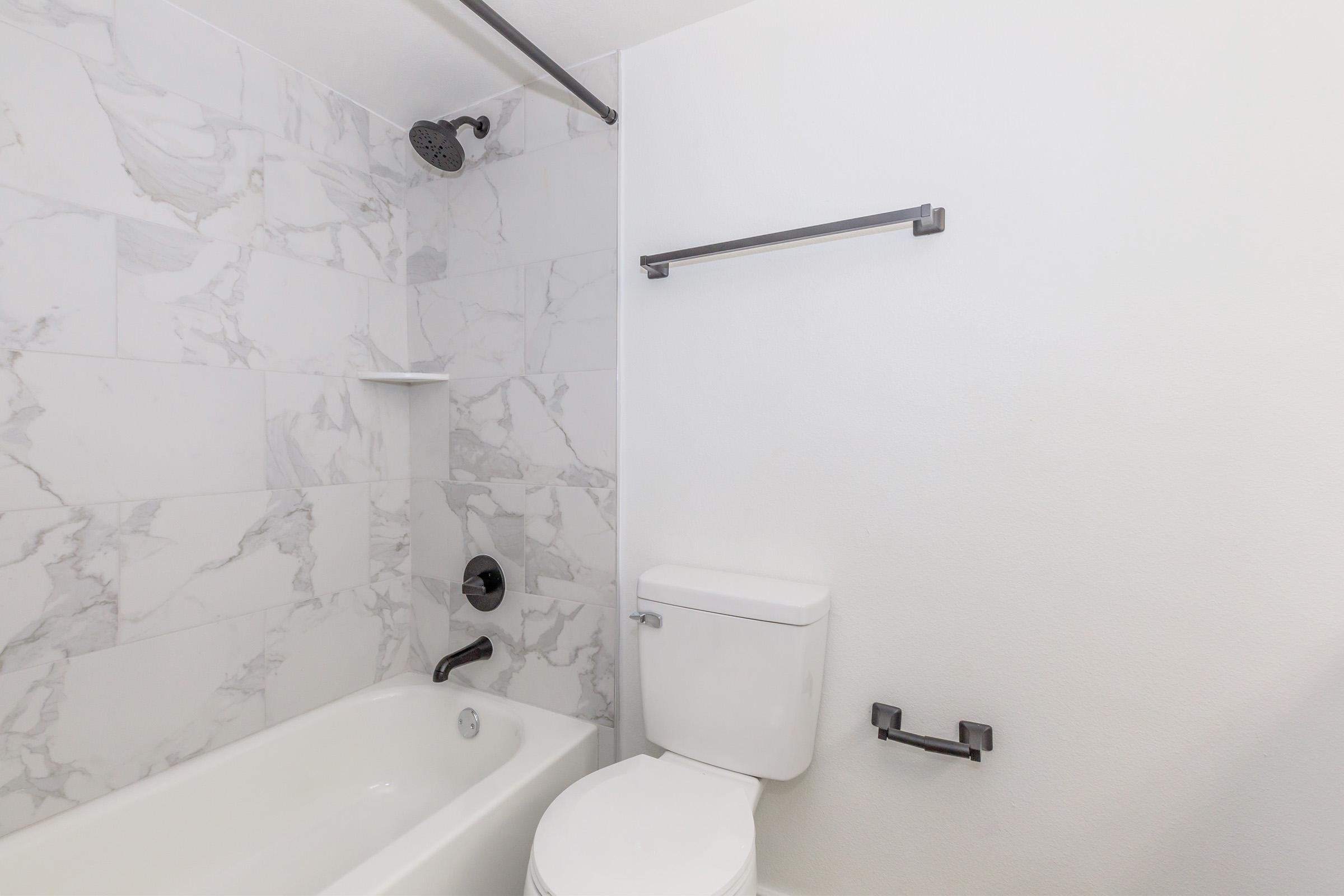 A modern bathroom featuring a bathtub with a showerhead, a white toilet, and a wall-mounted towel bar. The walls are covered in grey marble tiles, and the overall design is clean and minimalistic. The space is well-lit and uncluttered, creating a fresh and inviting atmosphere.
