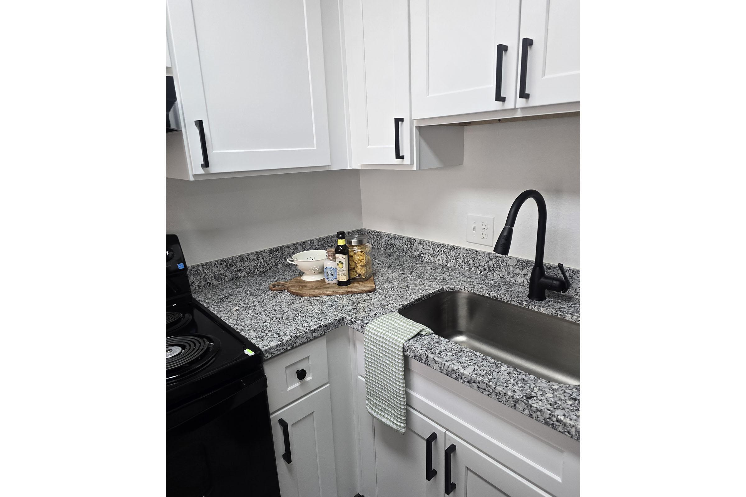 A modern kitchen corner featuring white cabinets with black handles, a granite countertop, a sink with a black faucet, and an electric stove. A wooden cutting board holds olive oil and spices, while a light green dish towel hangs from the cabinet edge.