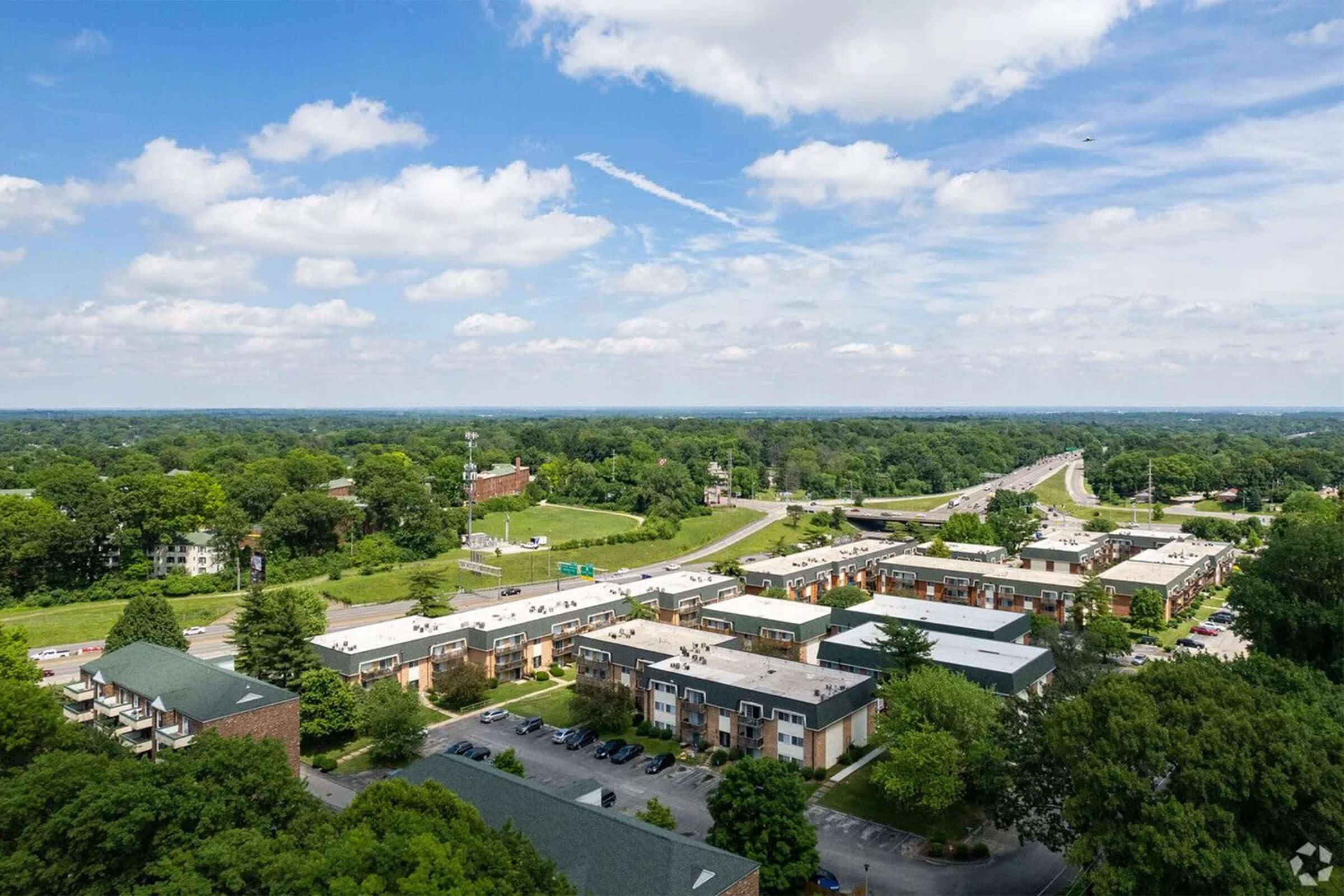 Aerial view of a suburban area featuring a mix of commercial buildings and residential apartments, bordered by green trees. A highway runs through the scene, with blue skies and scattered clouds overhead, highlighting the landscape's natural beauty and urban development.