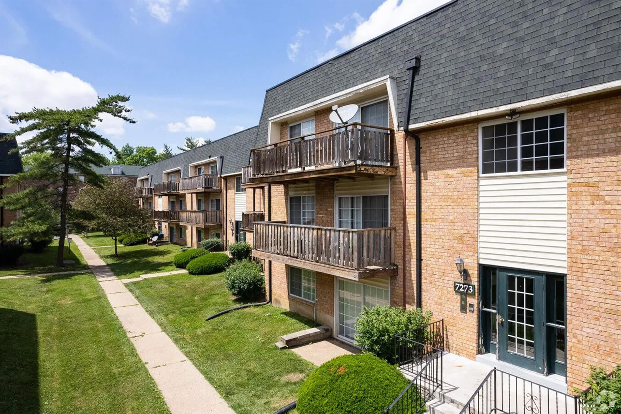 Brick apartment buildings with multiple balconies line a well-maintained pathway and landscaped green areas under a clear blue sky.