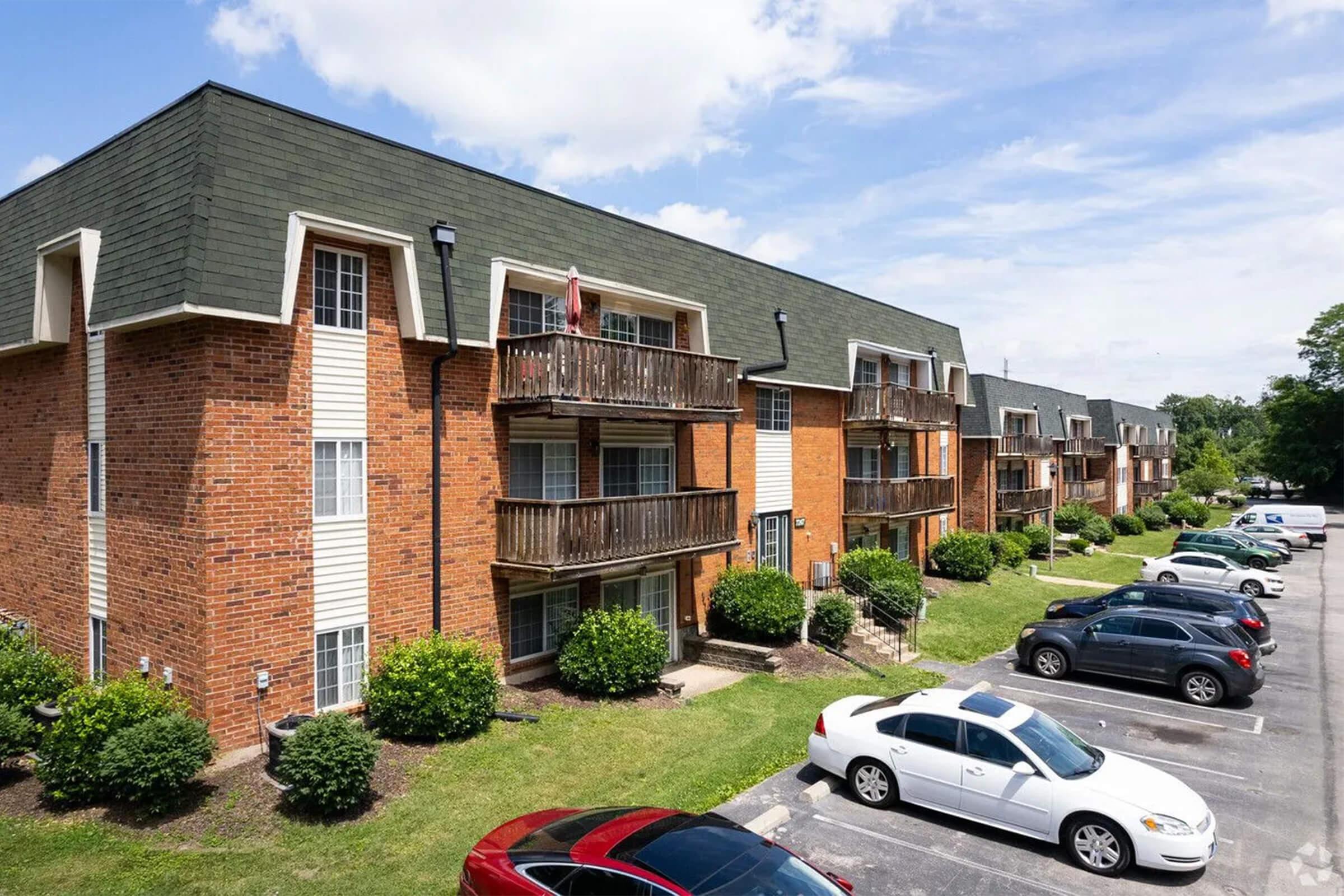 A row of brick apartment buildings with balconies, featuring green bushes in front and parked cars in the lot. The sky is partly cloudy, indicating a bright day. The architecture shows a modern design with a sloped roof.