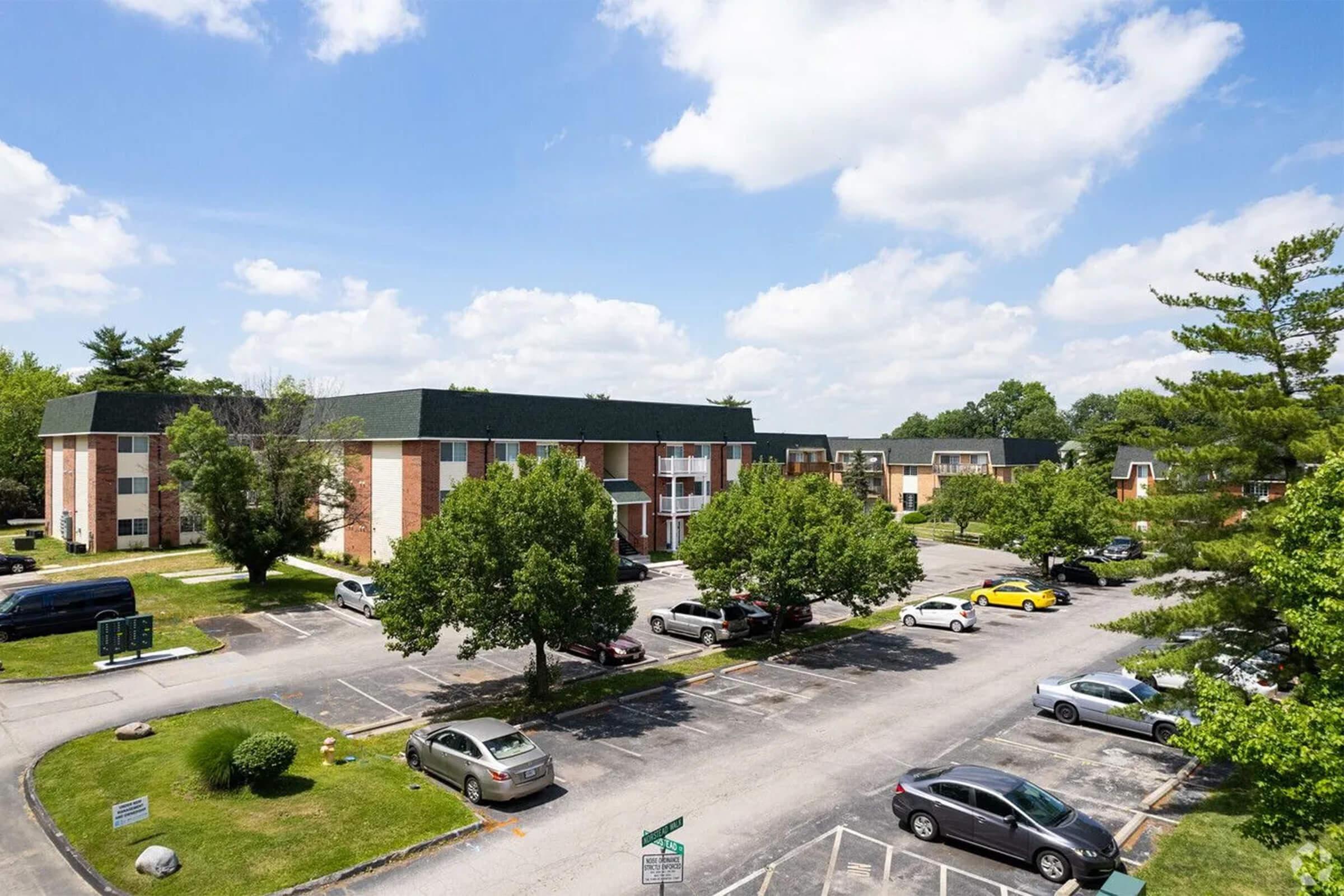 A view of a residential apartment complex featuring two-story buildings with brick and siding exteriors. The area includes a parking lot filled with cars and several trees providing greenery. The sky is partly cloudy, showcasing a pleasant day.