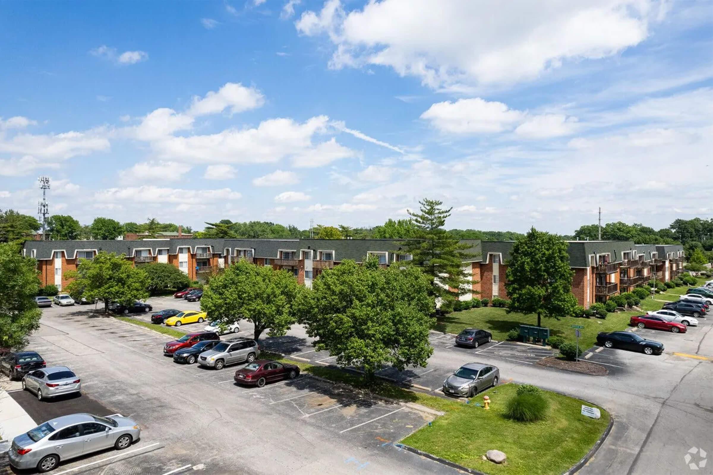 A wide view of a residential apartment complex, showcasing multiple two-story buildings surrounded by greenery. The parking lot in front is filled with various parked cars, and blue skies with scattered clouds are visible overhead.
