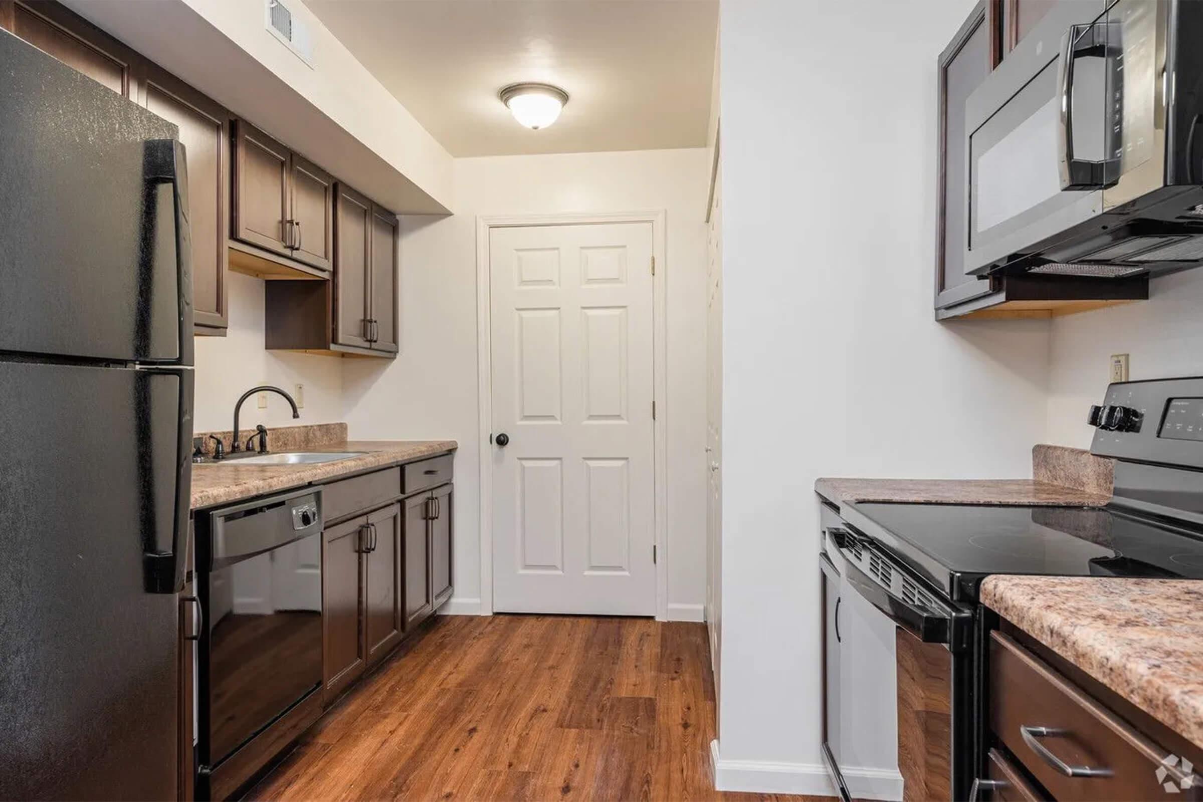 Modern kitchen featuring dark wood cabinetry, stainless steel appliances including a refrigerator, dishwasher, and microwave, along with a granite countertop. The space includes a door leading to another area and has laminate flooring for a contemporary look.