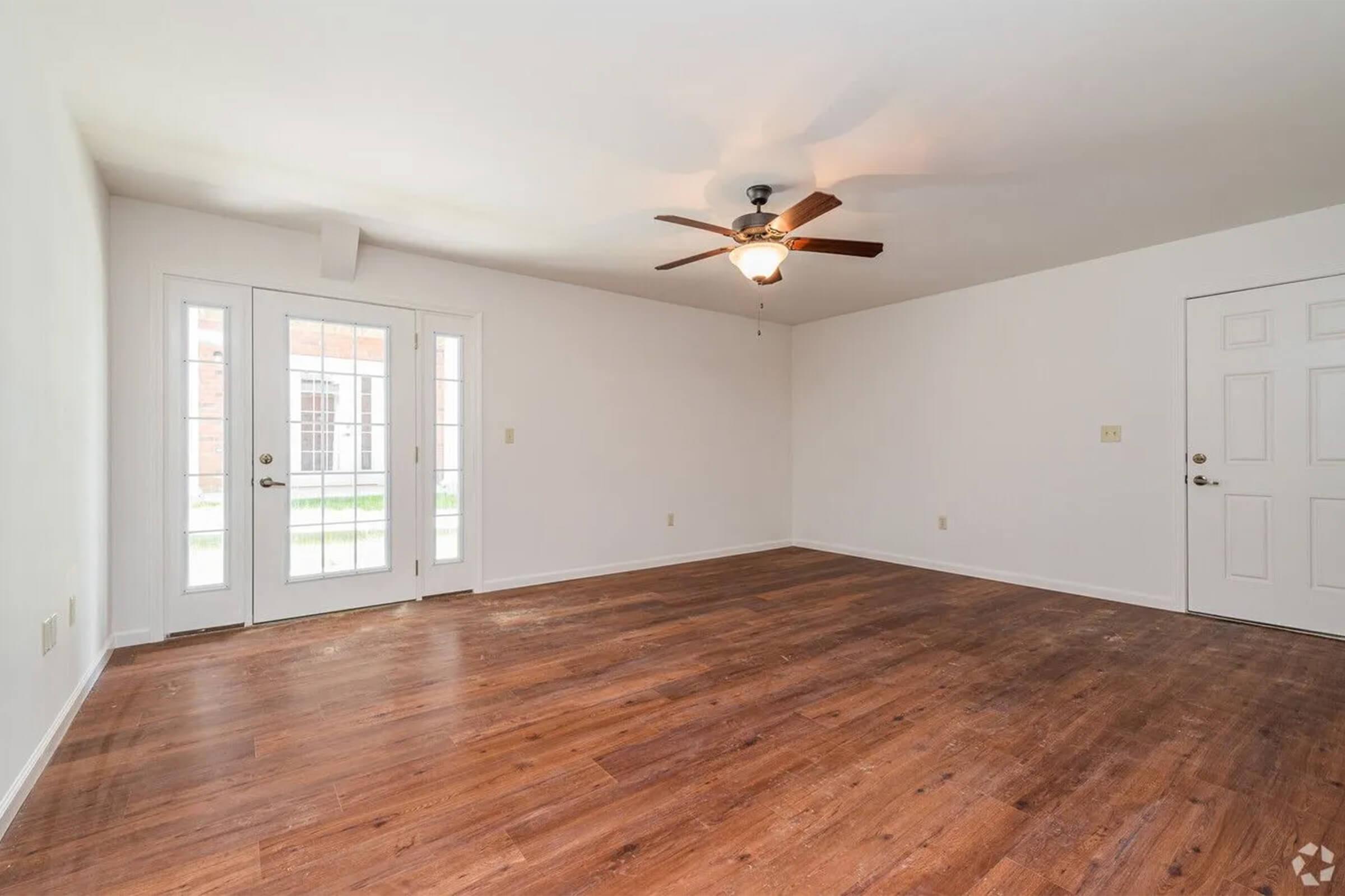 A spacious, empty room with light-colored walls and a wooden floor. A ceiling fan is mounted in the center, and there are two glass doors leading to an outdoor patio area. A door on the right side leads to another part of the home. Natural light fills the space, creating a bright and inviting atmosphere.