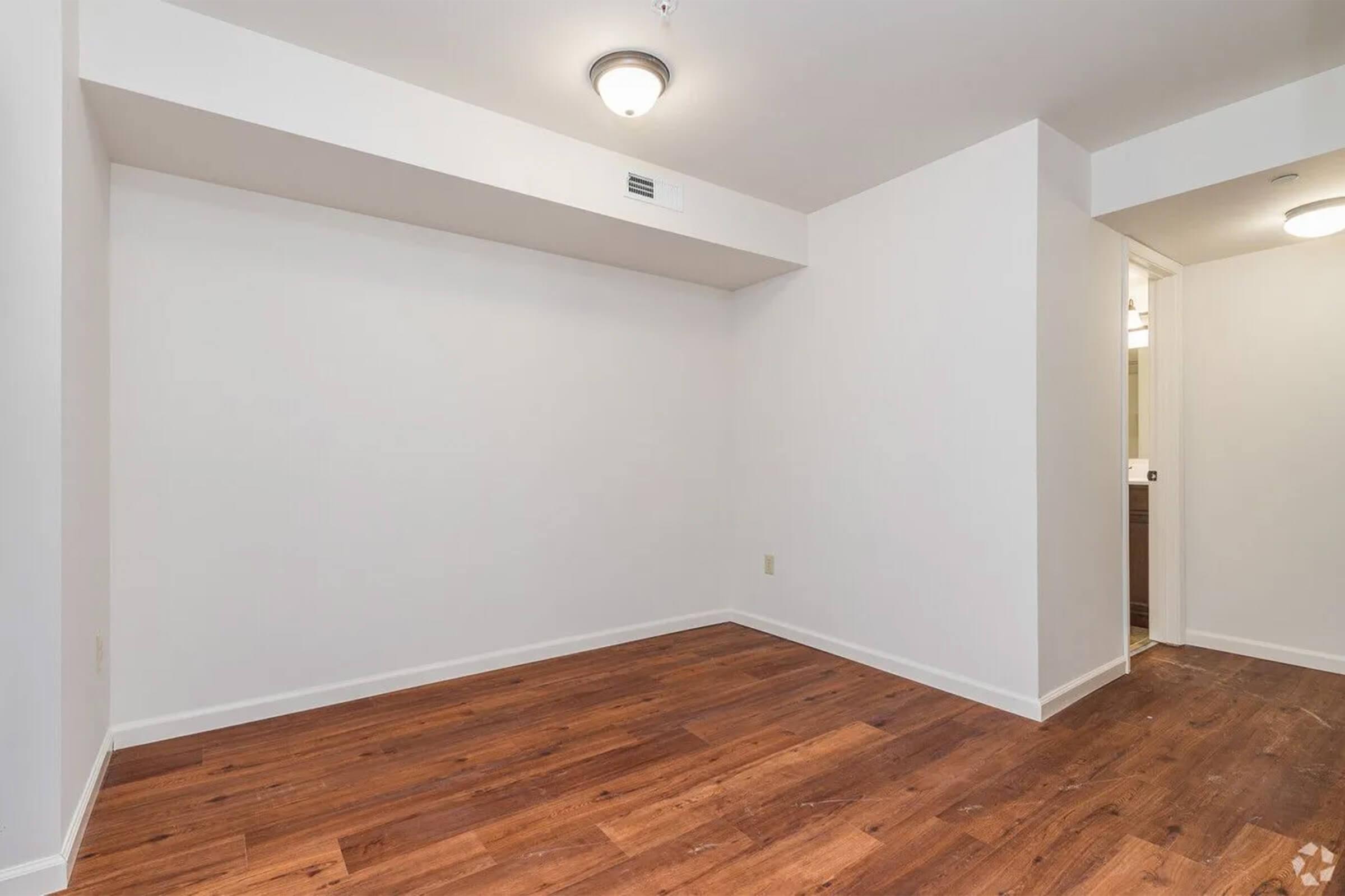 A sparse room with light-colored walls and a wooden floor. The space features a ceiling light fixture, and there is a doorway leading to a bathroom. The room is empty, suggesting it is ready for furniture or decoration.