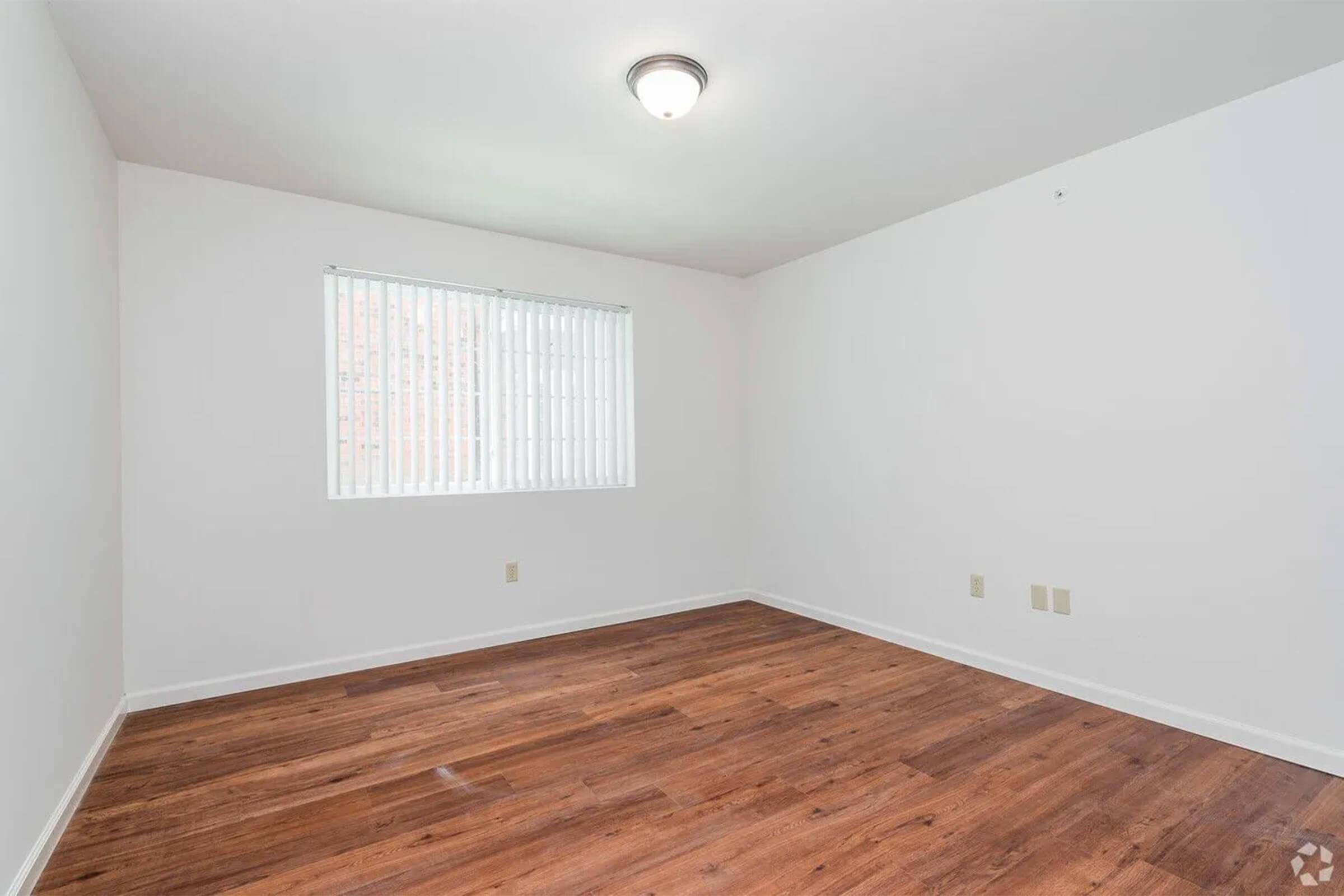 Empty room with light hardwood flooring, white walls, and a single window with vertical blinds letting in natural light. A ceiling light fixture is present, and there are electrical outlets on the walls. The overall atmosphere is bright and minimalistic.