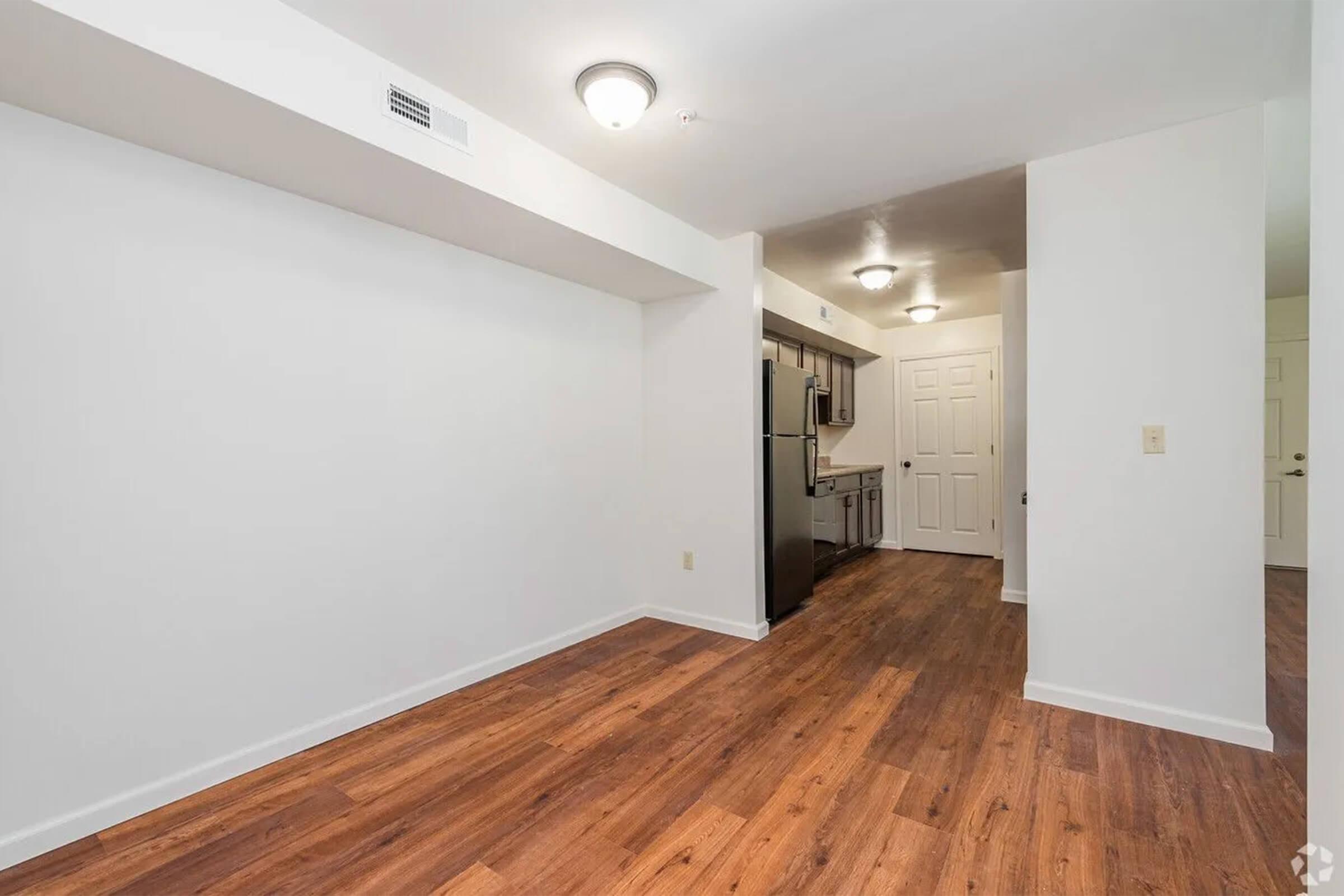 A spacious, modern living area featuring a light-colored wall with a wood floor. On one side, there's a kitchen equipped with stainless steel appliances, including a refrigerator. The room is well-lit with ceiling lights, creating a bright and inviting atmosphere.