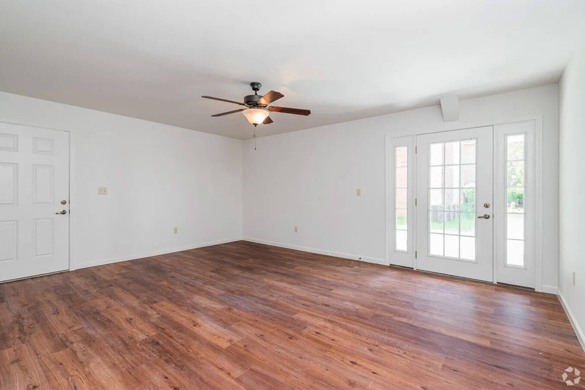 An empty living room featuring light-colored walls and wood-like flooring. A ceiling fan with light fixture hangs from the ceiling, and there are two doors visible on one wall, leading to an outdoor area. Natural light enters through the glass doors, creating a bright and open atmosphere.