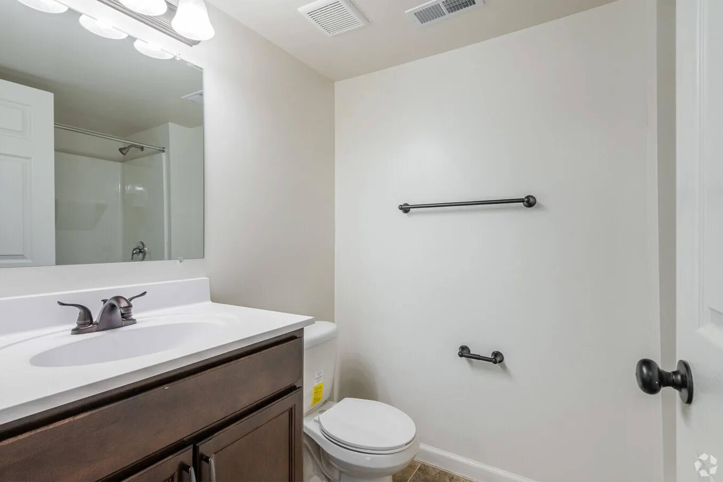 A clean bathroom featuring a white sink with a dark wood cabinet, a toilet, and a towel rack. The walls are light-colored, and there is a shower area visible in the background. Bright lighting from a ceiling fixture adds to the spacious feel of the room.