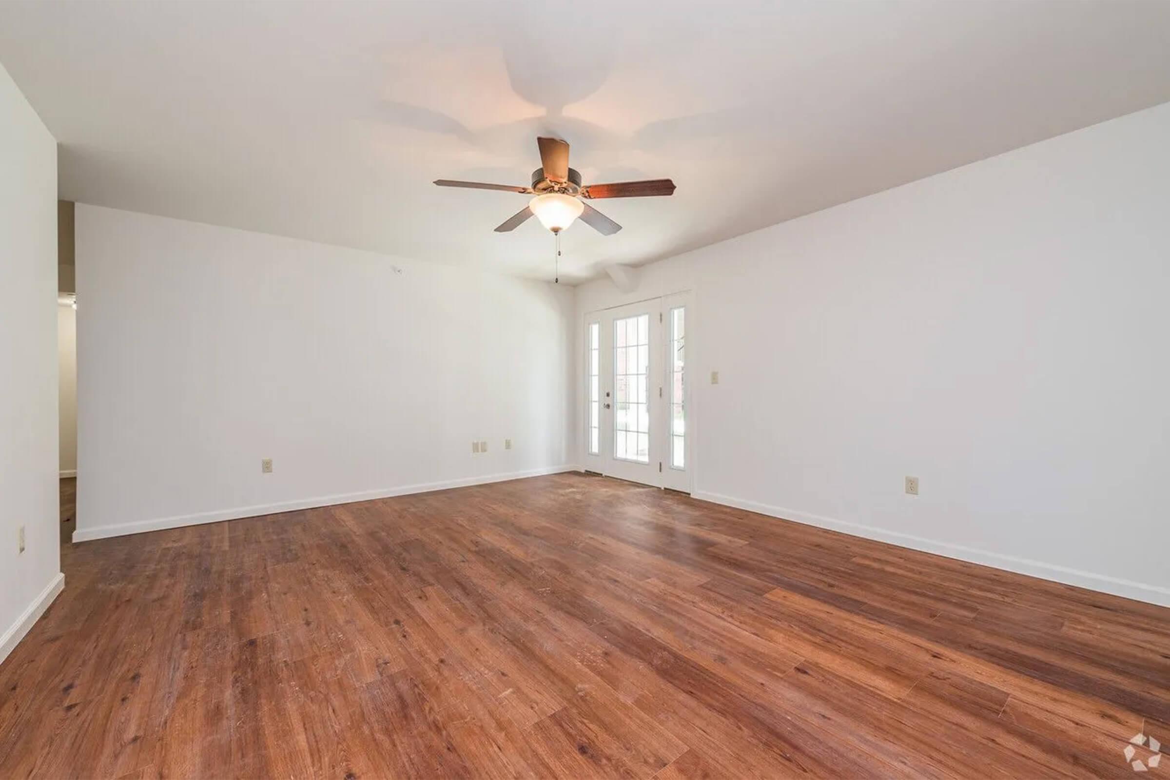 A spacious, empty living room with white walls and a ceiling fan. It features a hardwood floor, large windows, and a set of French doors leading outside. The room is well-lit and has no furniture, creating a minimalist and inviting atmosphere.