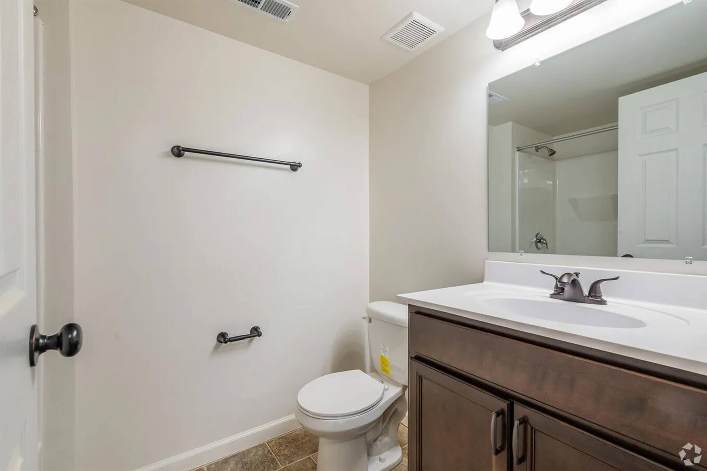 A clean, modern bathroom featuring a toilet, a single sink vanity with a dark wood cabinet, a large mirror, and a towel rack. The walls are painted in a neutral color, and there is a shower visible in the background. The floor has a tile finish.