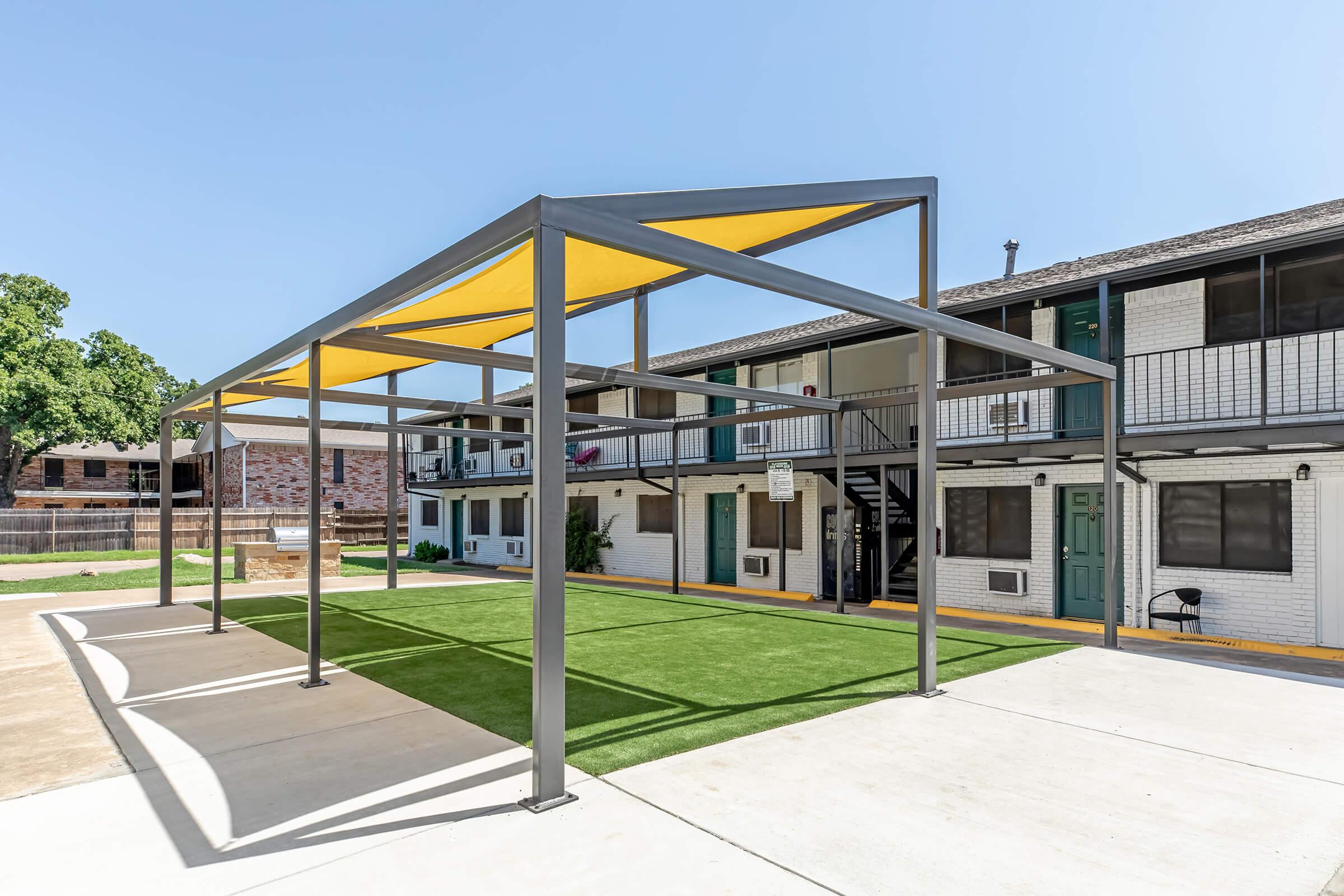 A steel frame structure with a yellow canopy provides shade over a green turf area in front of a multi-story building. The building features multiple doors with a staircase on one side, and the setting is sunny with a clear blue sky.