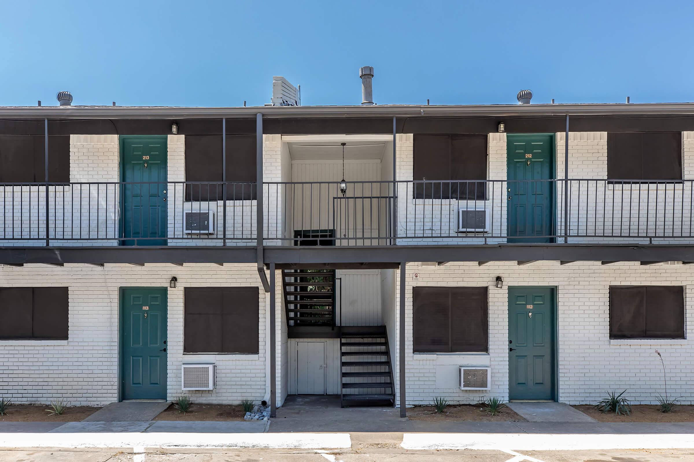 Two-story residential building with a white brick exterior, featuring blue-green doors and black window coverings. A central staircase leads to the upper level, and there are air conditioning units visible beneath the windows. The ground is gravel with minimal landscaping.