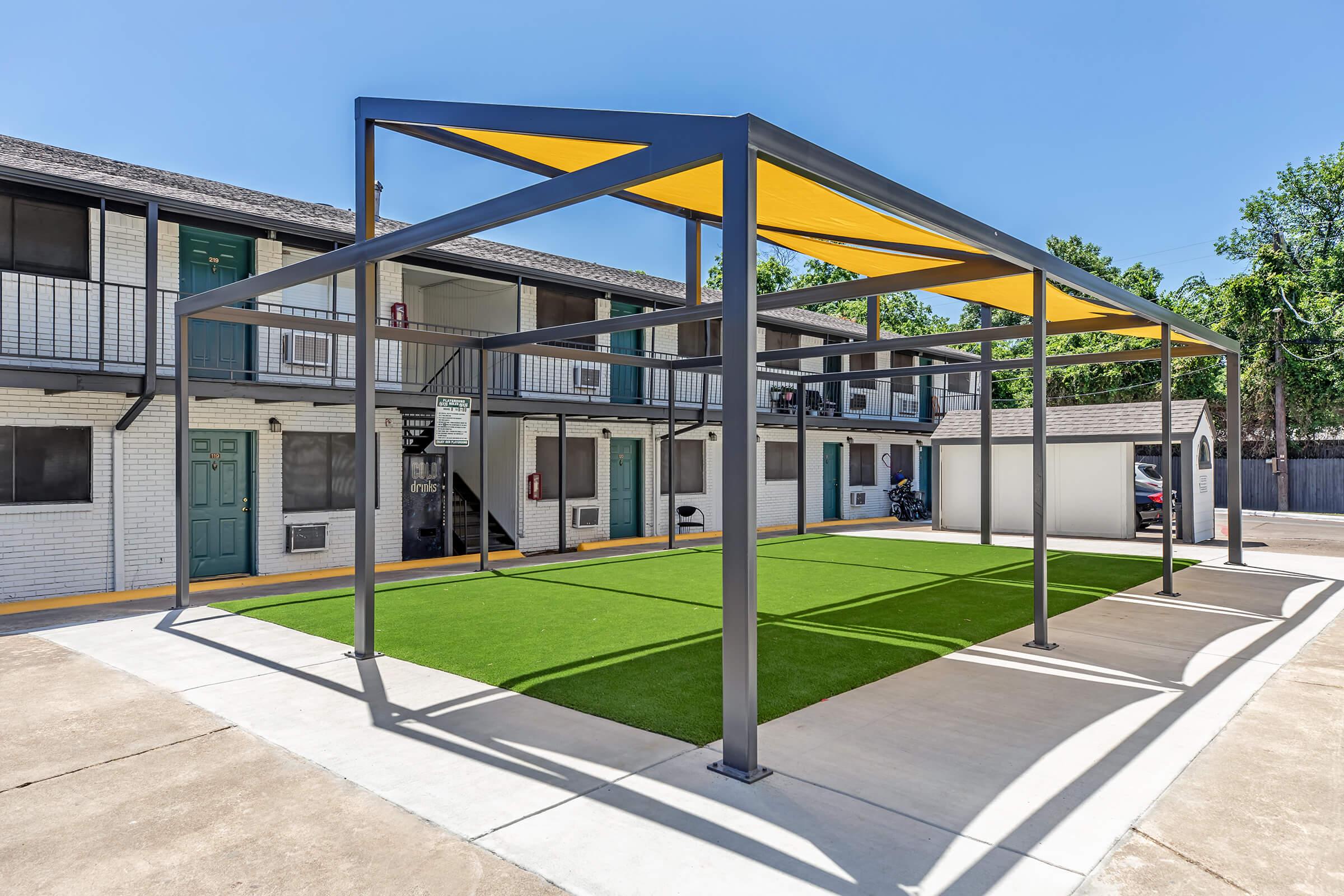 A modern outdoor area featuring a green artificial turf space sheltered by a yellow canopy supported by gray metal beams. Surrounding the area are two-story buildings with white walls and balconies, along with parking spaces visible in the background. Clear blue sky overhead adds to the inviting atmosphere.