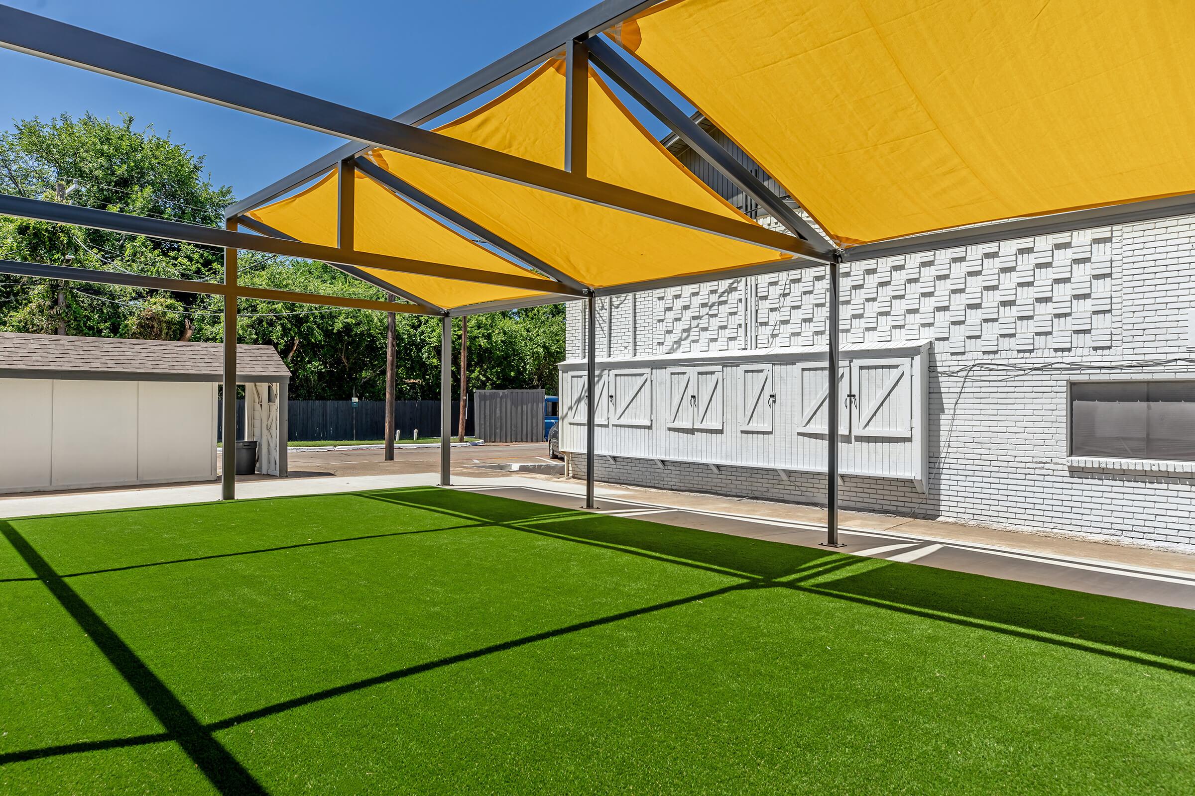 A spacious outdoor area featuring artificial turf and a vibrant yellow canopy structure. In the background, a white building is visible along with a storage shed. The scene is well-lit, showcasing a clear blue sky and greenery around the perimeter.