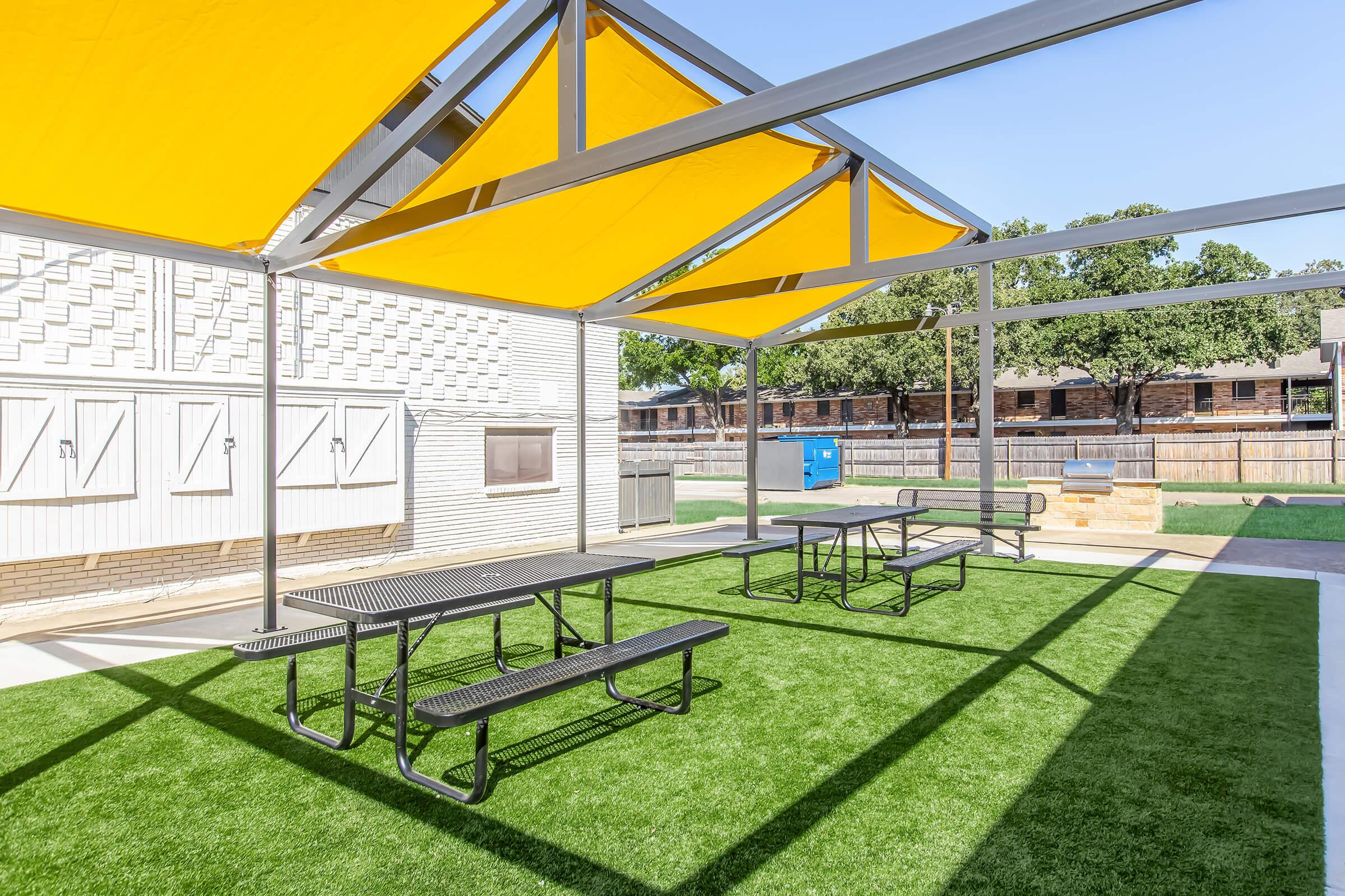 Outdoor picnic area featuring several black metal tables and benches under bright yellow canopies. The space has artificial turf and is surrounded by trees and nearby buildings, providing a pleasant environment for gatherings or meals.