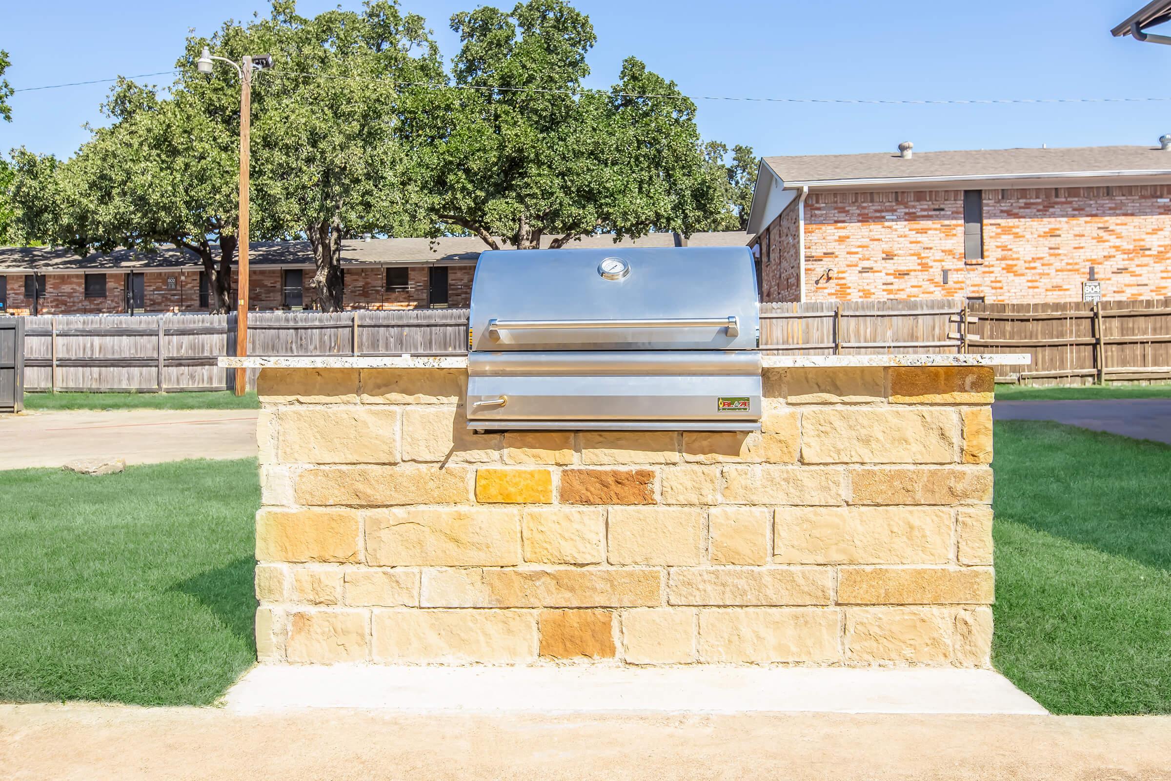 A stainless steel grill mounted on a stone wall, set in a grassy backyard. In the background, there are residential buildings with a wooden fence and trees lining the area. The sky is clear and blue, indicating a sunny day.