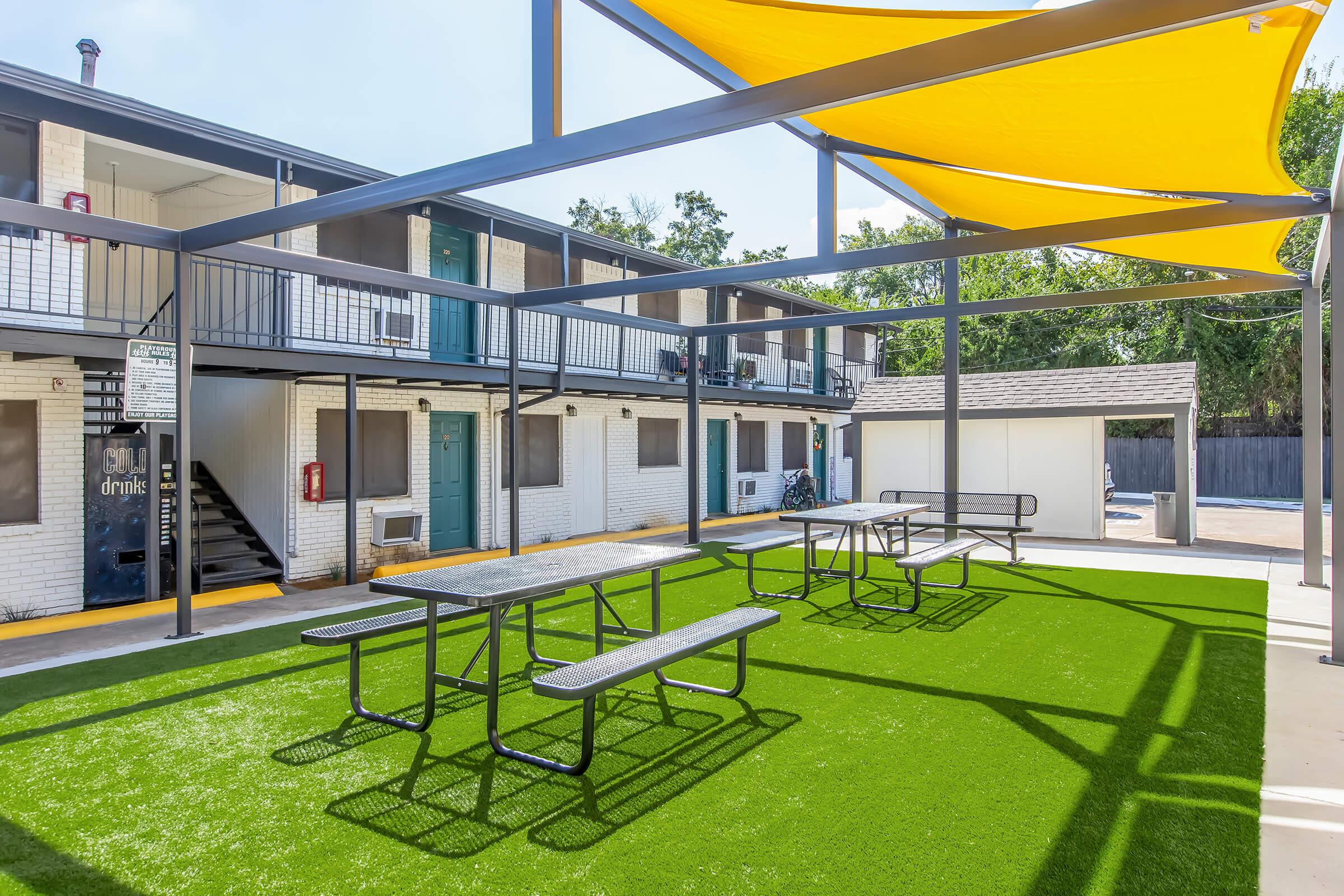 Outdoor shaded area with multiple picnic tables on green turf, surrounded by a two-story building featuring several white rooms and air conditioning units. A small structure is visible in the background, and the scene is set under clear blue skies.