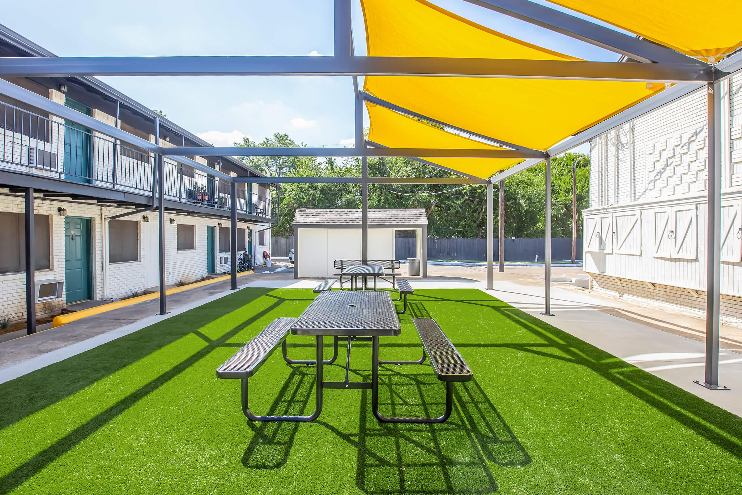 An outdoor area featuring a picnic table under a large yellow shade, surrounded by green artificial turf. The setting includes adjacent buildings and a small storage shed in the background, with clear skies above.