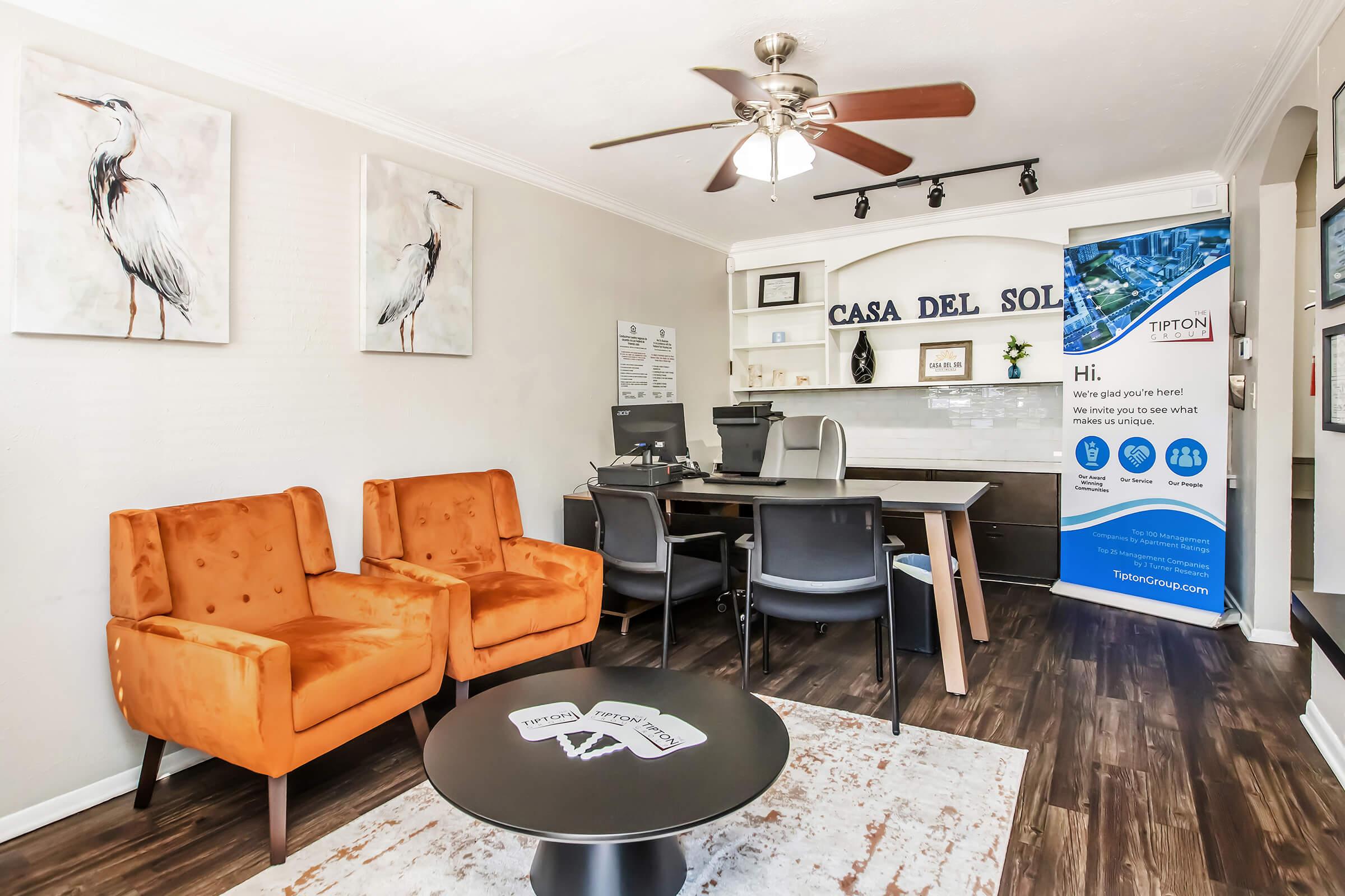 Cozy office space featuring two orange armchairs, a modern black coffee table, and a sleek desk. The walls are decorated with bird-themed artwork, and there is a banner displaying the Tipton Group logo. A ceiling fan adds to the inviting atmosphere.