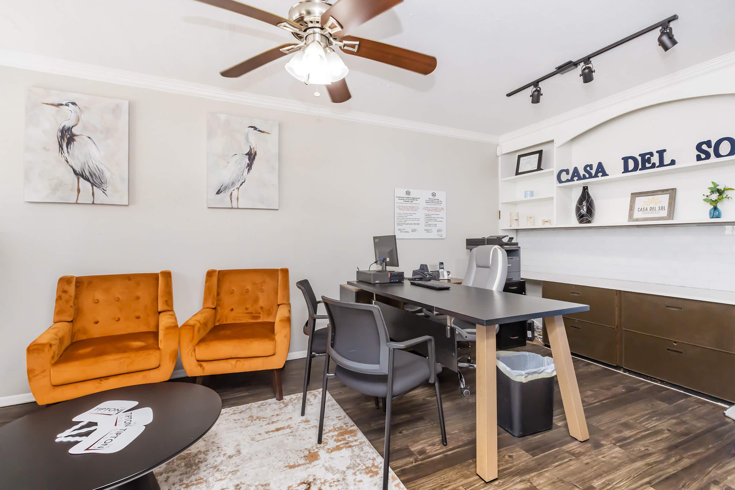 A modern office space featuring two orange armchairs and a sleek black desk with a computer. The walls are decorated with framed bird artwork, and there are shelves with decorative items. A small round coffee table is visible in front, and a wastebasket is placed nearby. Natural light enhances the inviting atmosphere.