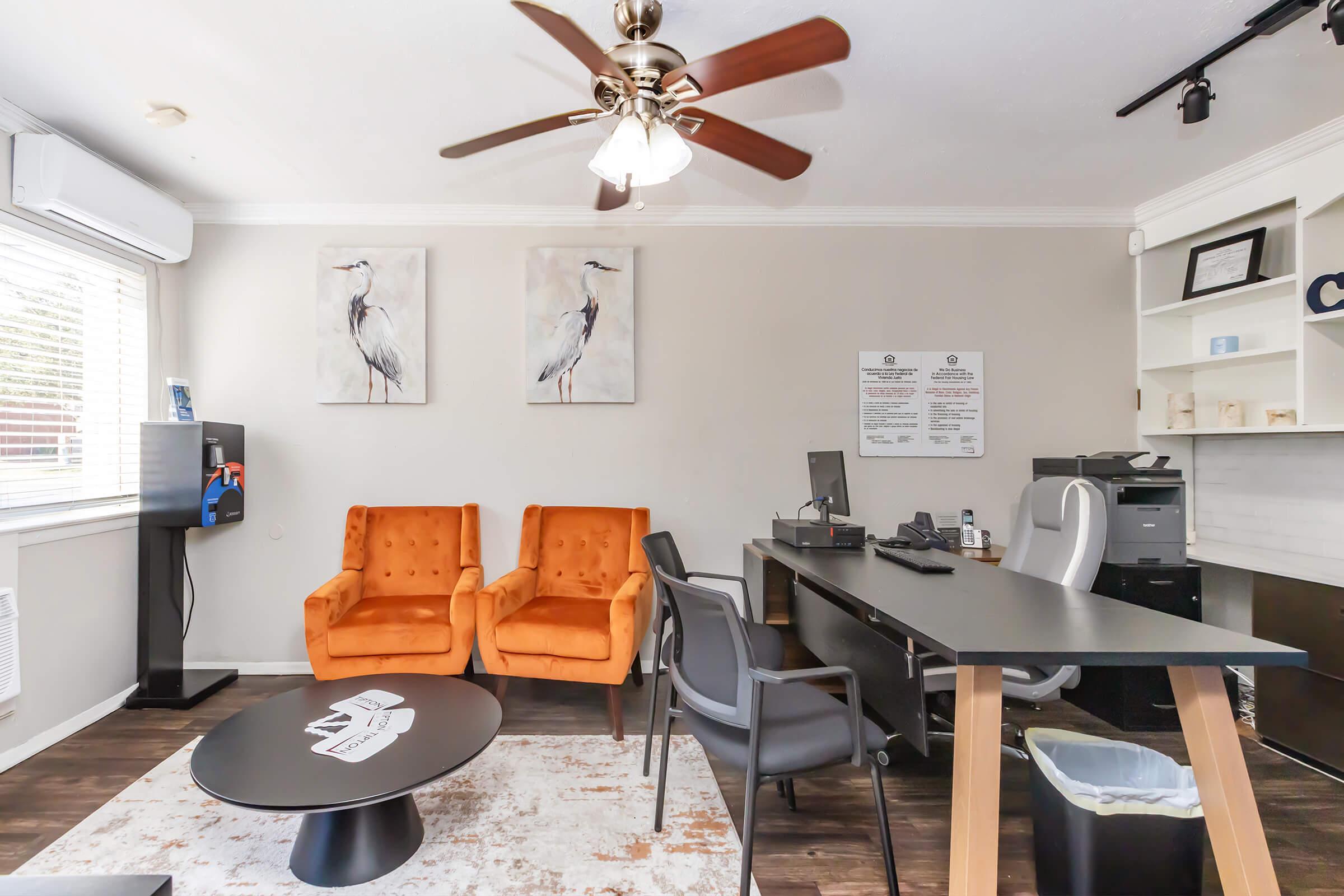 A modern office space featuring two bright orange armchairs, a black desk with a computer, a printer, and a document holder. There is a round black coffee table in the center, a fan overhead, and wall art depicting herons. Natural light comes through a window, enhancing the inviting atmosphere.