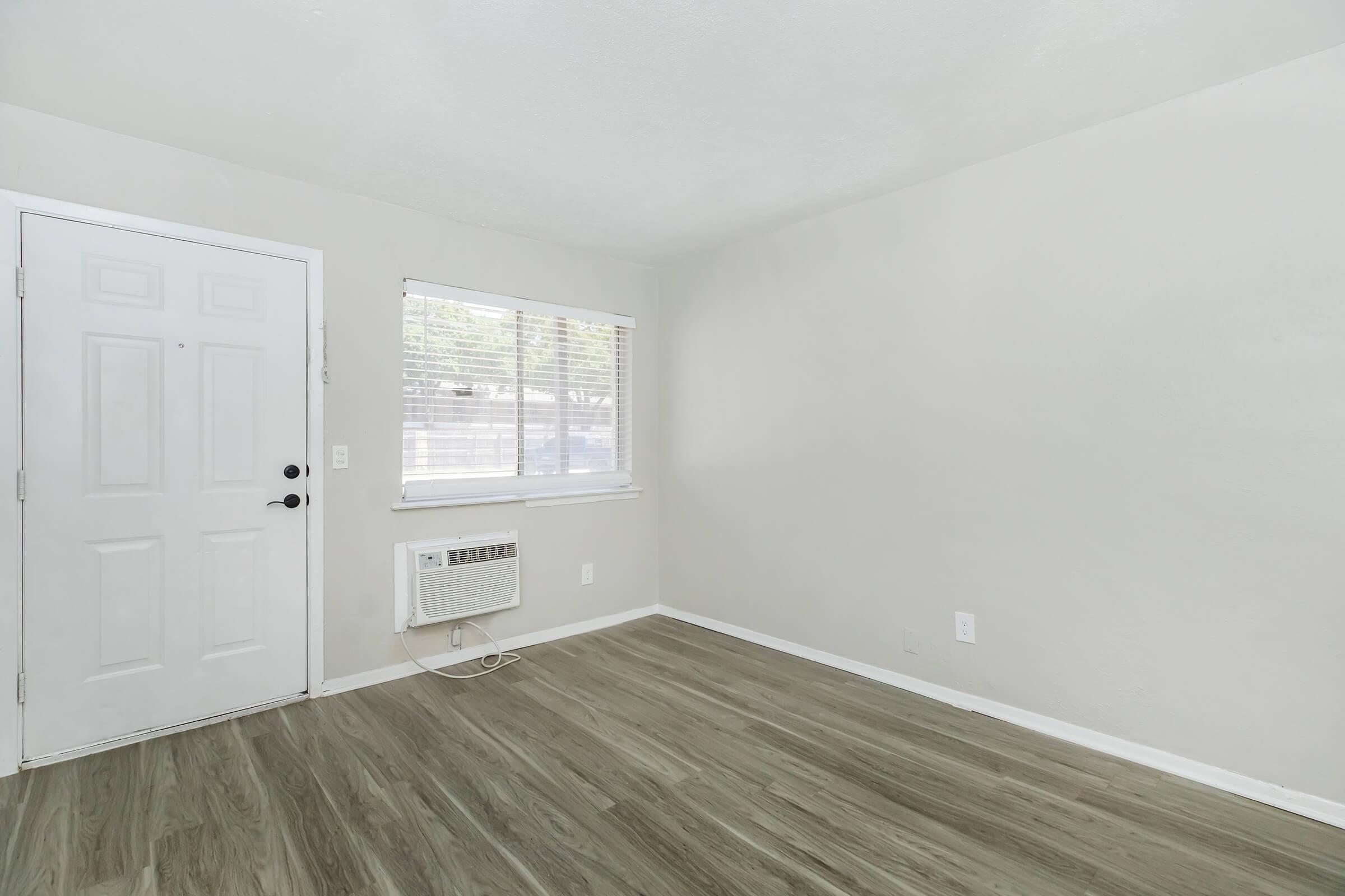 A well-lit, empty room with light gray walls, a vinyl floor, and a window covered with blinds. There is an air conditioning unit mounted on the wall beneath the window. A white door is visible on the left side of the image.