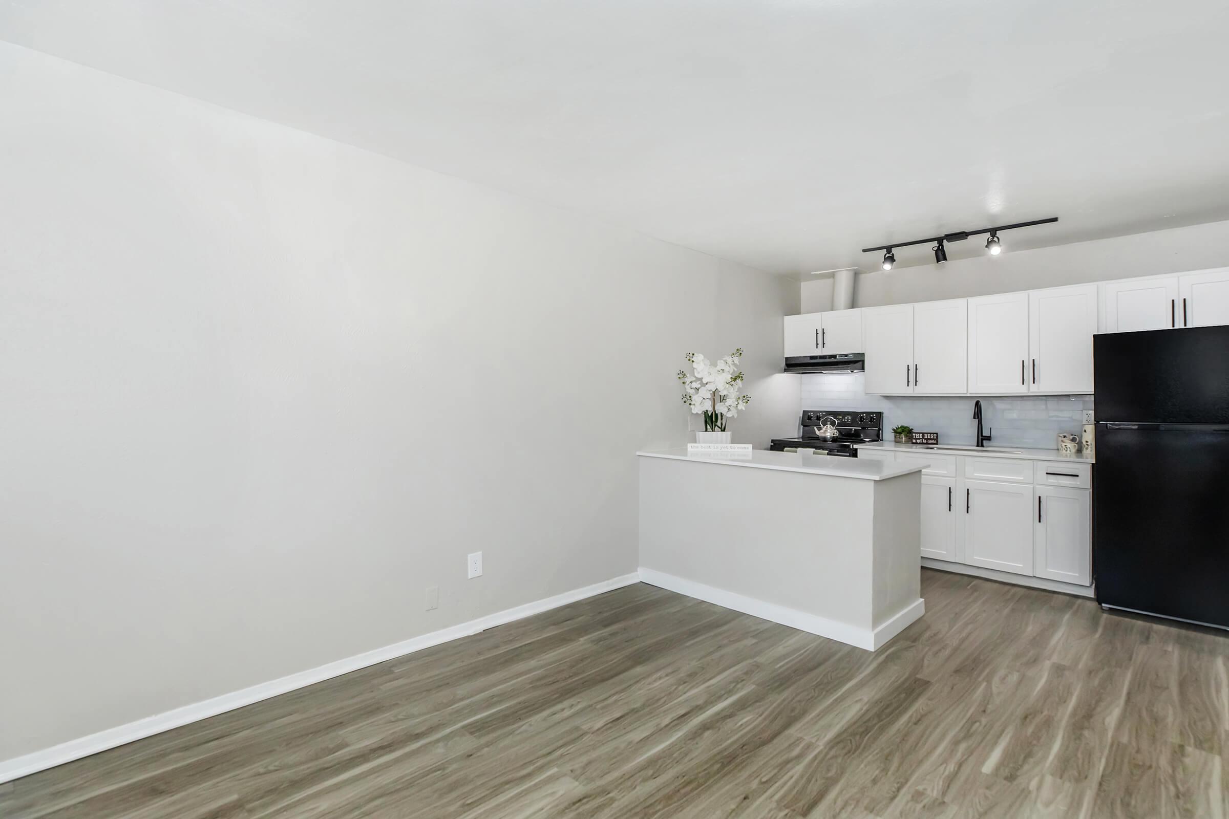 A modern, spacious apartment interior featuring a light color scheme. An open kitchen area with white cabinets, black appliances, and a small island. The floor has a wood-like finish, and there are decorative flowers on the counter. The walls are plain and light-colored, creating a bright and airy atmosphere.