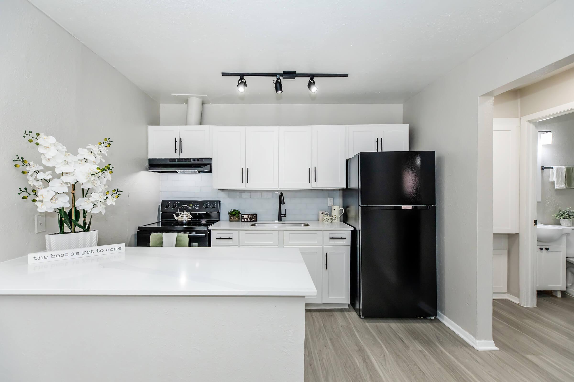 A modern kitchen featuring white cabinetry, a black refrigerator, and an electric stove. The countertops are light-colored with a decorative sign stating "the best is yet to come." Elegant white orchids add a touch of decor. The space has light gray walls and wood-like flooring, enhancing its contemporary look.