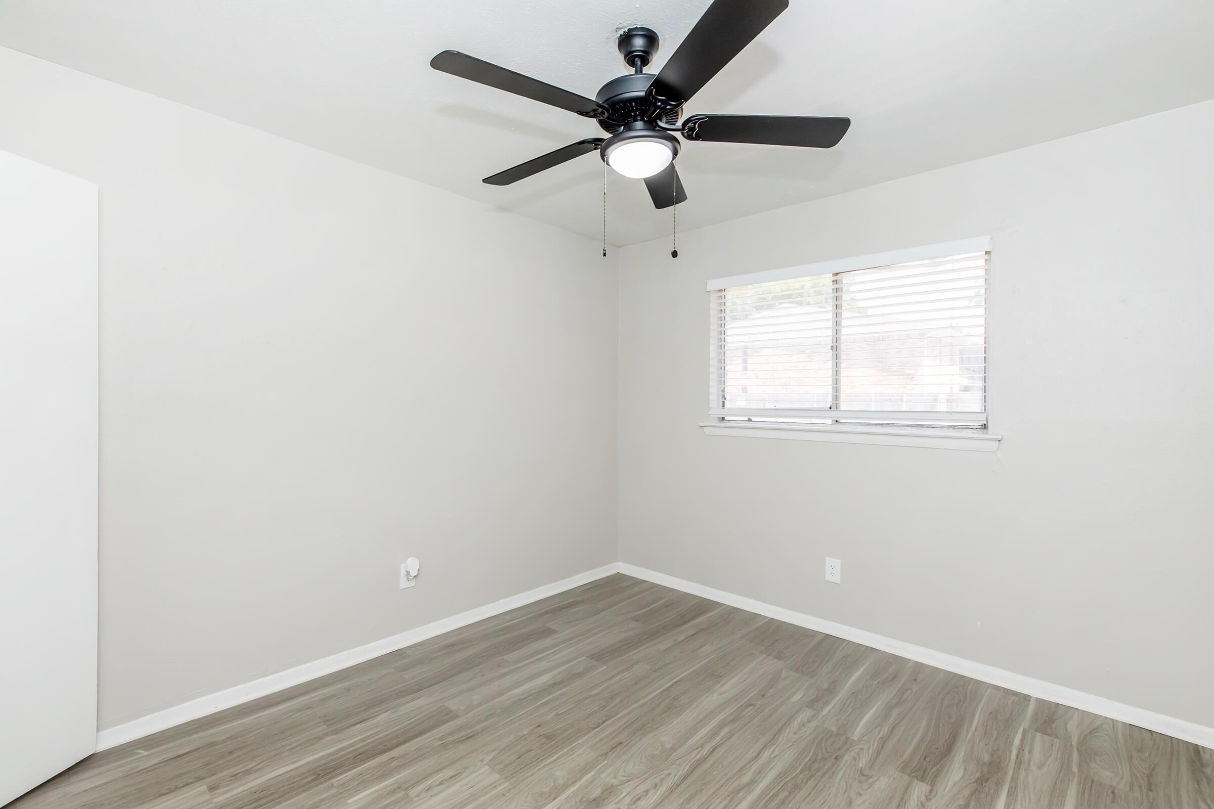 A clean, empty room with light gray walls and a wooden floor. A ceiling fan with a light fixture is mounted in the center, and there is a window covered with blinds allowing natural light in. A white closet door is partially visible on the left side.