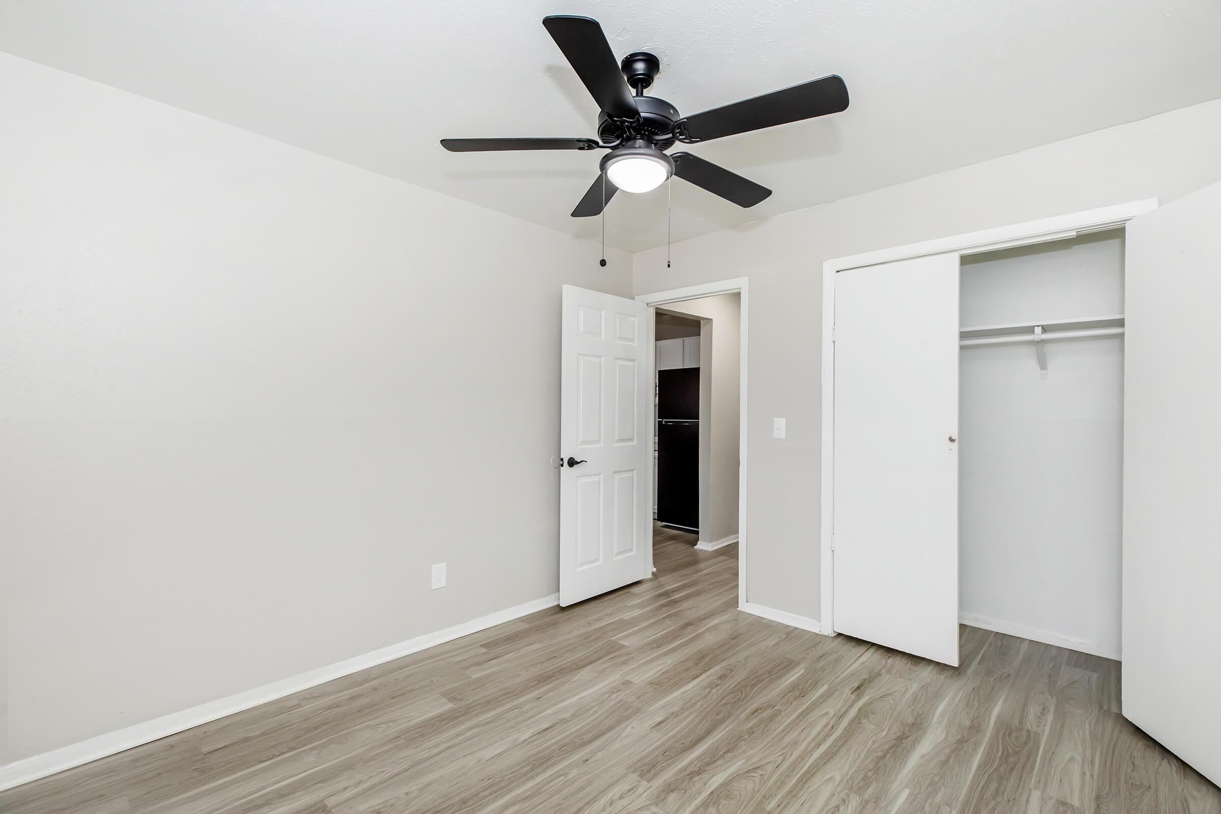 A bright, unfurnished bedroom featuring light-colored walls, a ceiling fan with a light fixture, and light wood flooring. The room includes a closet with double doors and an open doorway leading to another space, enhancing the airy and spacious feel of the area.