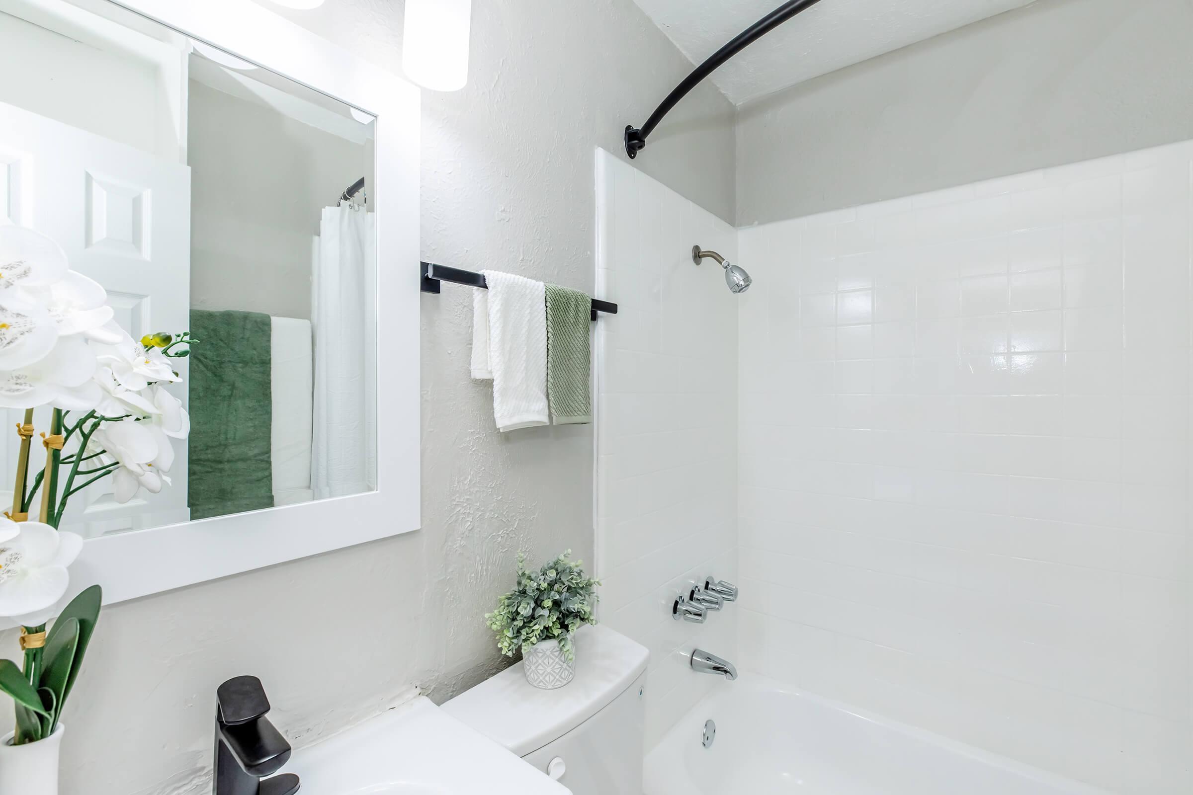 A clean and modern bathroom featuring a white tub with a showerhead, a large mirror, and a sink. Neatly hung towels in green and white are on a rack, with a small potted plant and a vase of white orchids adding decorative touches. The walls are painted in neutral tones for a bright appearance.