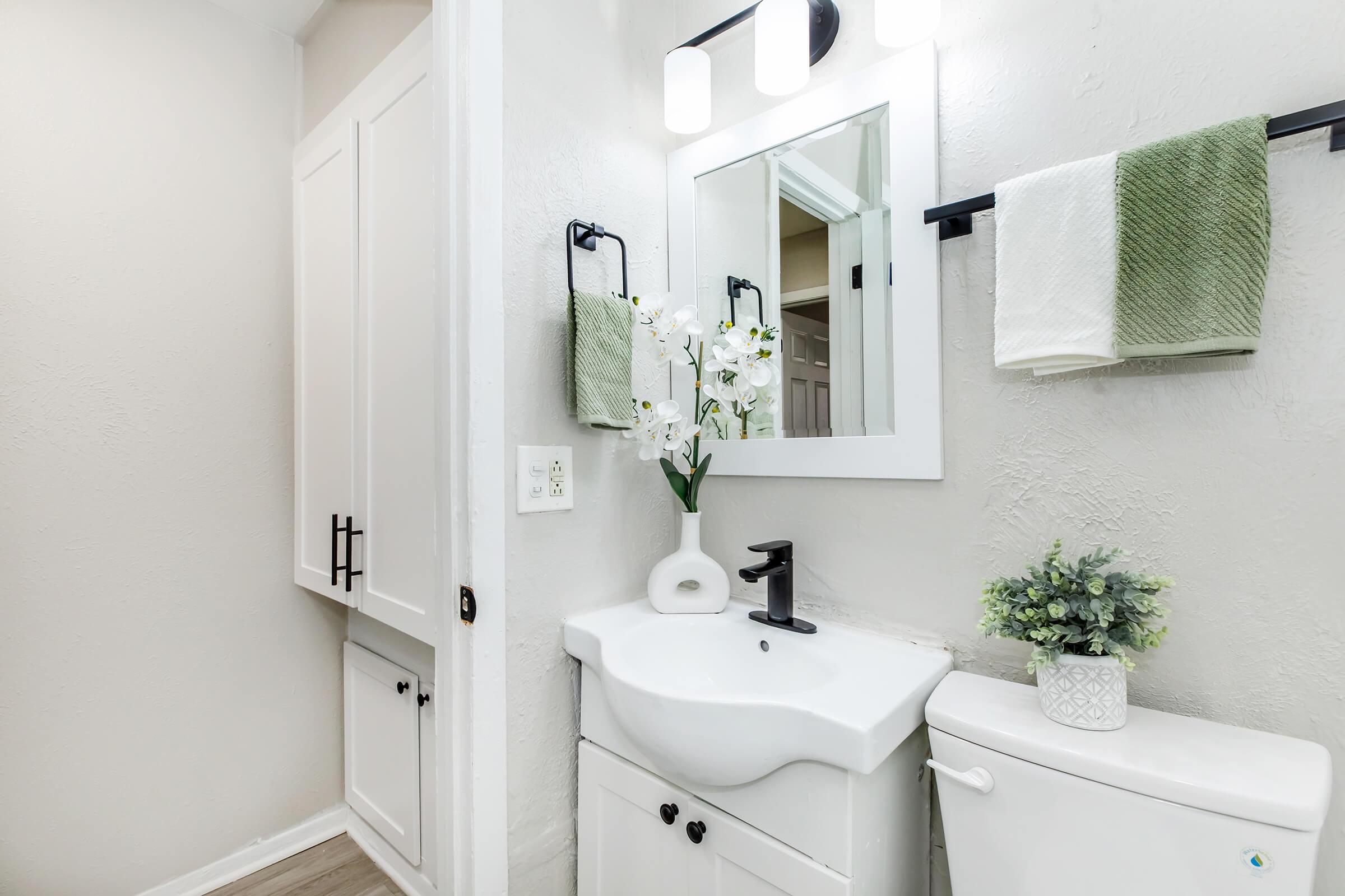 A modern bathroom featuring a white vanity with a sink, a mirror above, and decorative flowers. There's a small potted plant, a towel rack with green and white towels, and light-colored walls. The floor is a light wood tone, creating a clean and contemporary look.
