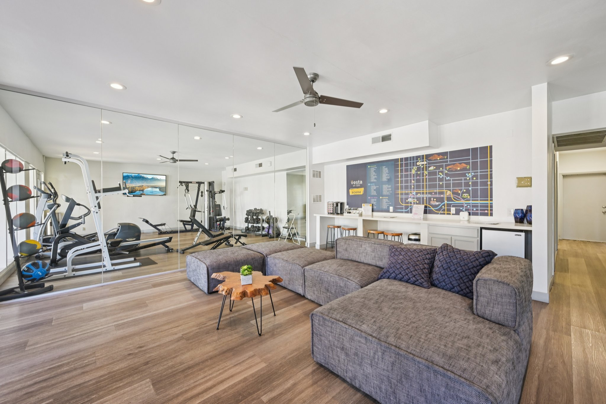 A modern, spacious living area featuring a grey sectional sofa with decorative pillows, a small wooden table, and a fitness area with exercise equipment visible in the background. The walls are bright, with mirrors and a large wall-mounted map or chart. Natural light fills the space through overhead lights.