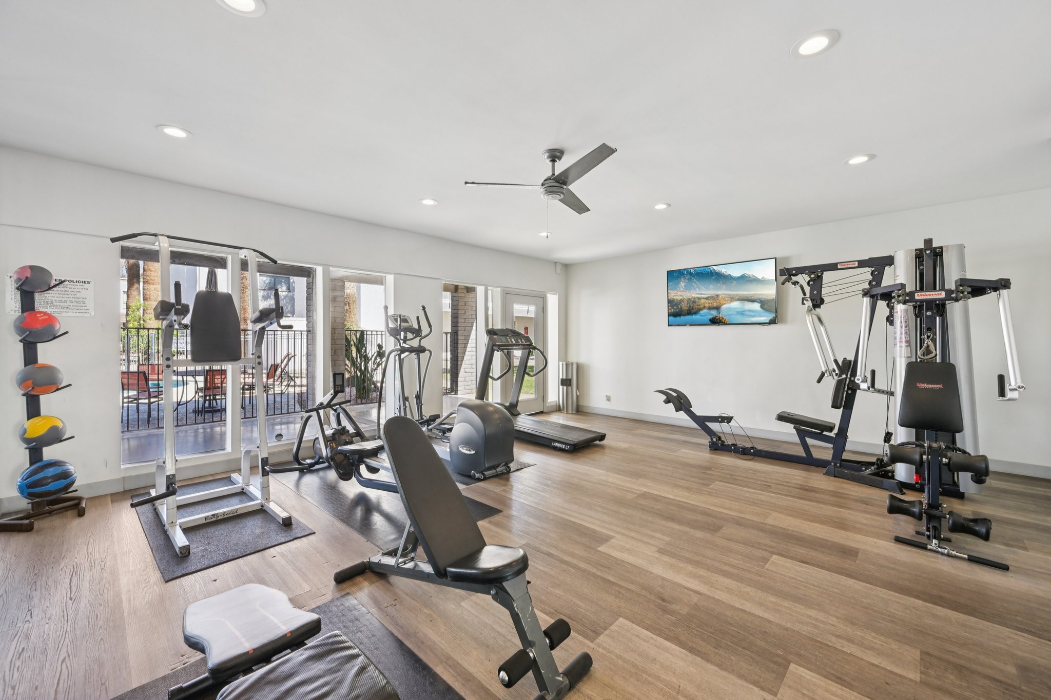 A modern gym interior featuring various exercise equipment, including a weight bench, treadmill, elliptical, and multi-gym. There are also exercise mats, a ceiling fan, and bright lighting. A large TV mounted on the wall displays a scenic mountain view. Windows provide natural light and a view of an outdoor space.