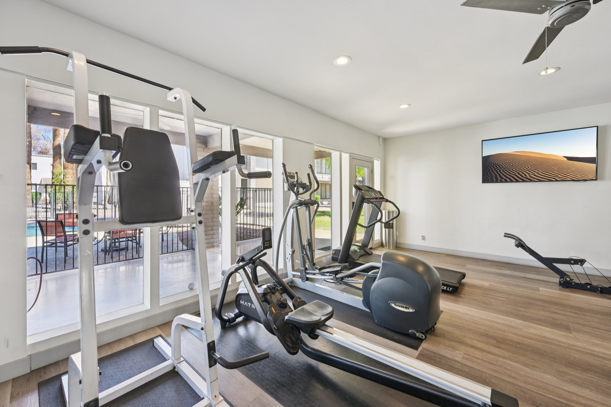 A modern gym interior featuring various fitness equipment, including an elliptical machine, a stationary bike, and a multi-gym machine. Large windows provide natural light and a view of an outdoor pool area. A flat-screen television mounted on the wall displays a serene desert landscape.
