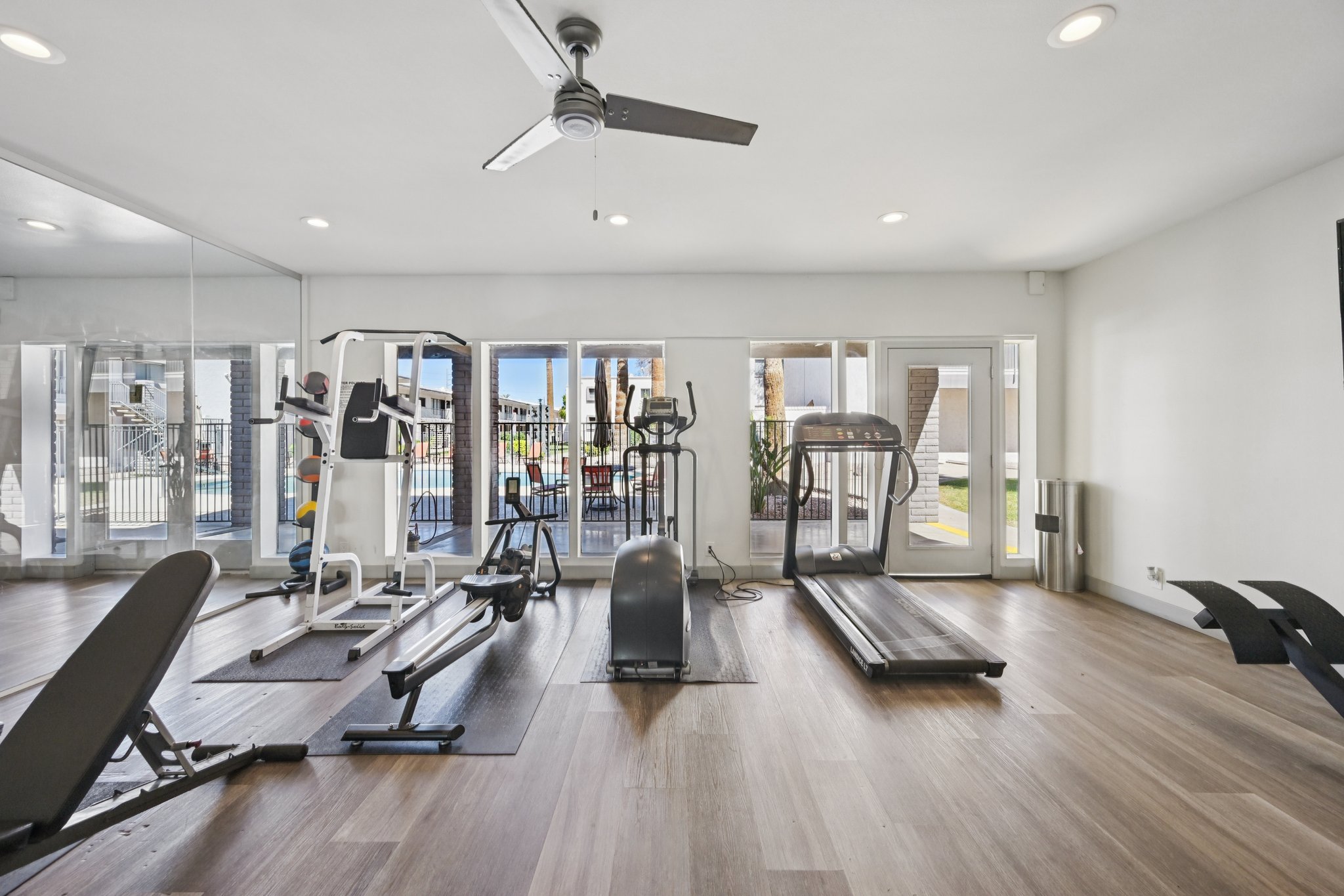 A modern gym interior featuring various exercise equipment, including treadmills, stationary bikes, and weight machines. Large windows provide natural light and a view of an outdoor area. The flooring is hardwood, and a ceiling fan is visible.